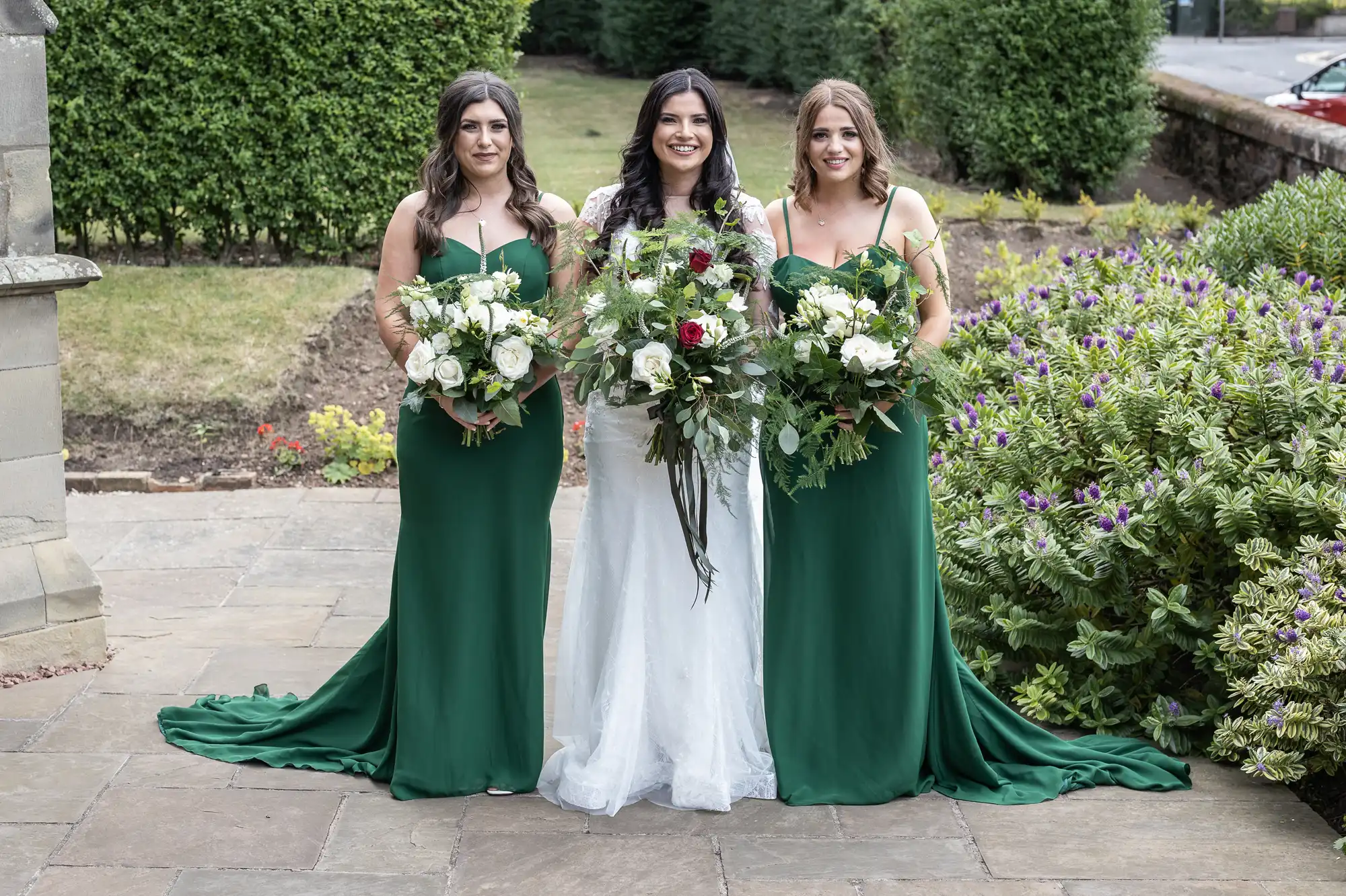 Three women in formal attire holding bouquets, two in green dresses and one in a white wedding dress, standing outdoors.