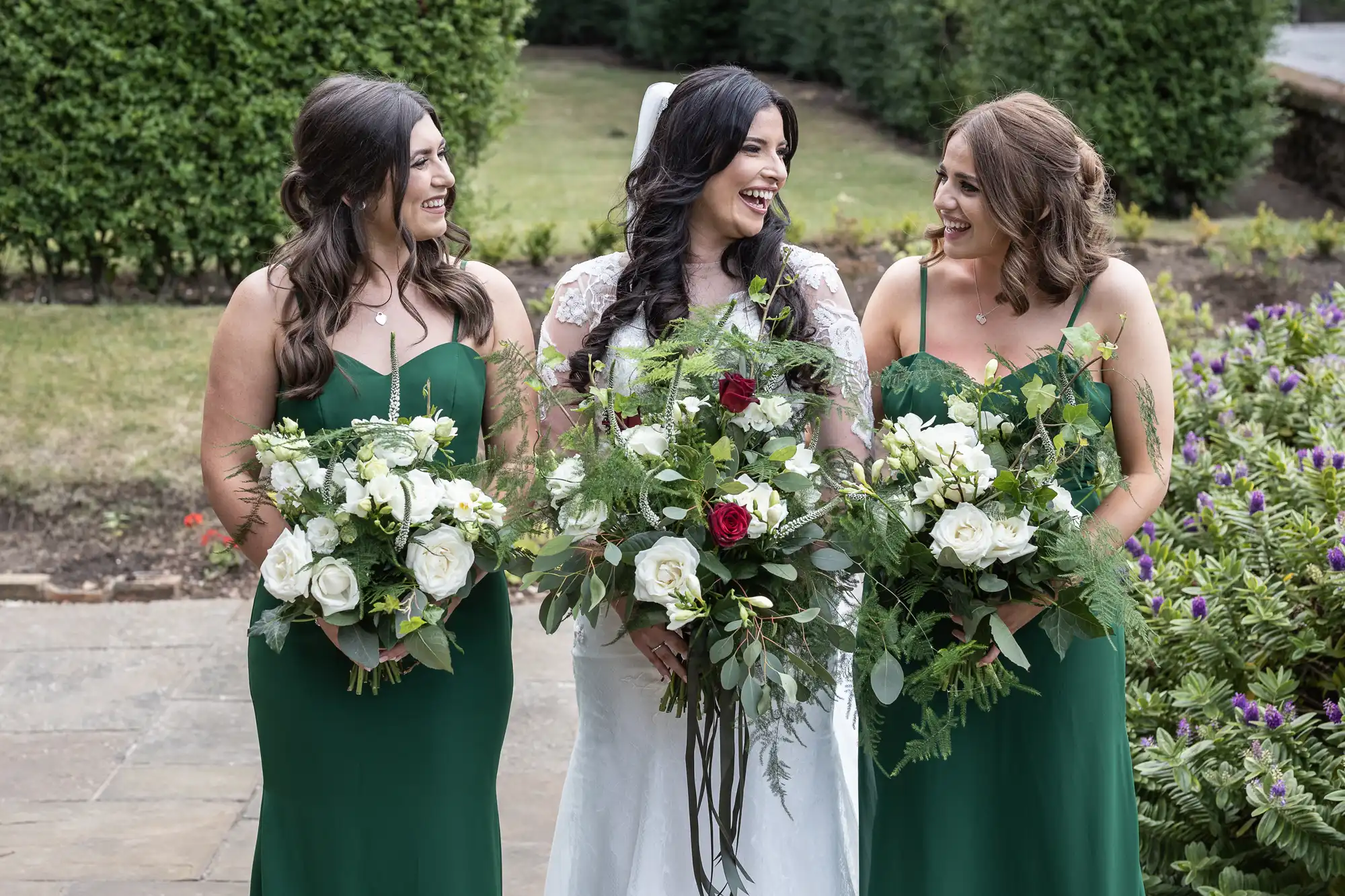 A bride in a white dress and two bridesmaids in green dresses, all holding floral bouquets, smiling and chatting outdoors.
