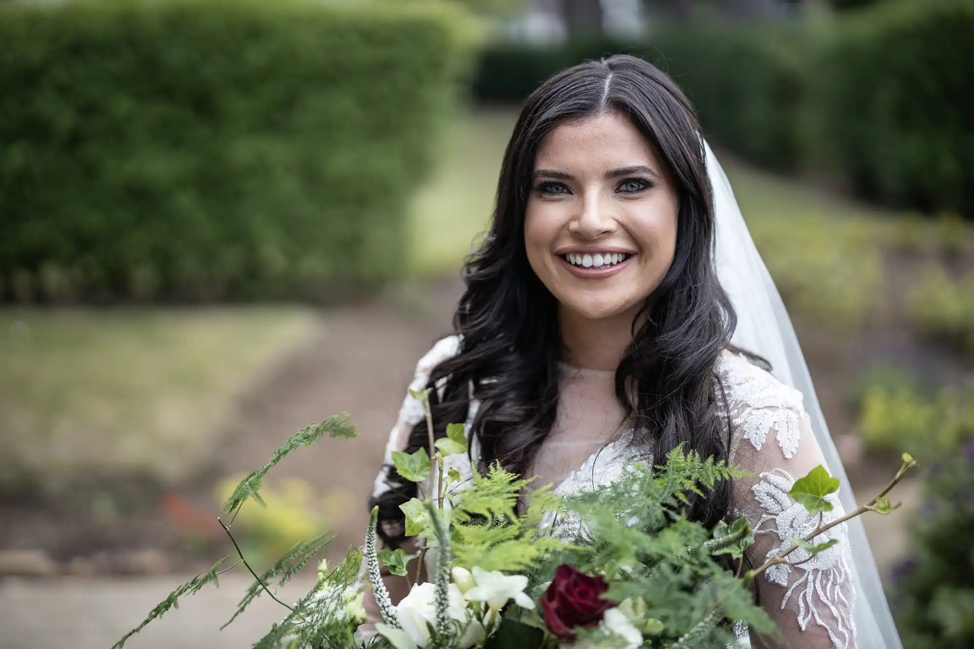 A radiant bride in a lace gown and veil, holding a bouquet, smiles joyously in a garden.