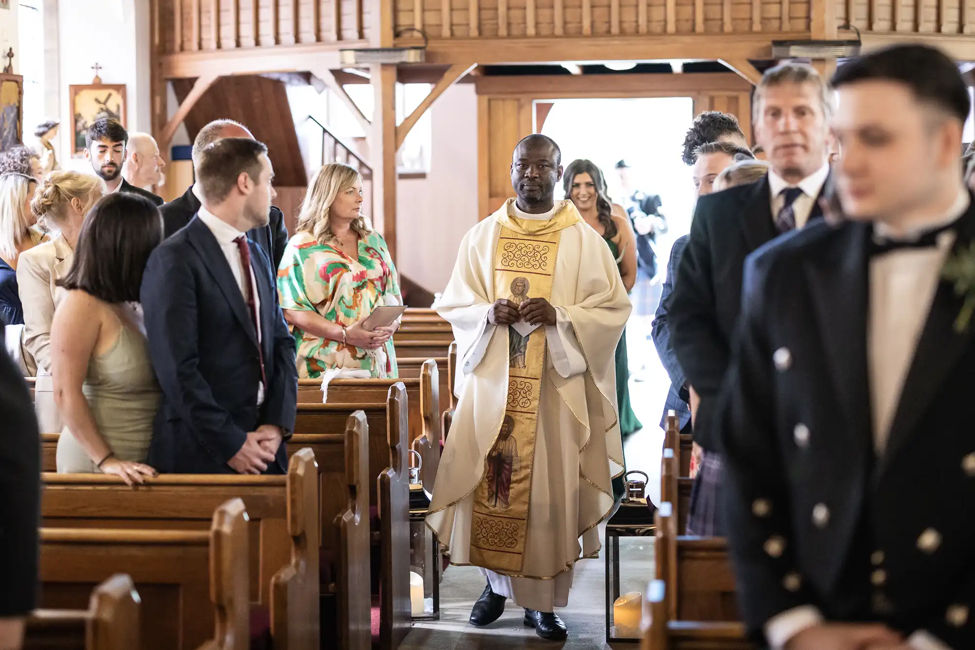 A priest walking down the aisle of a church during a wedding ceremony, with guests turning to look as he passes by.