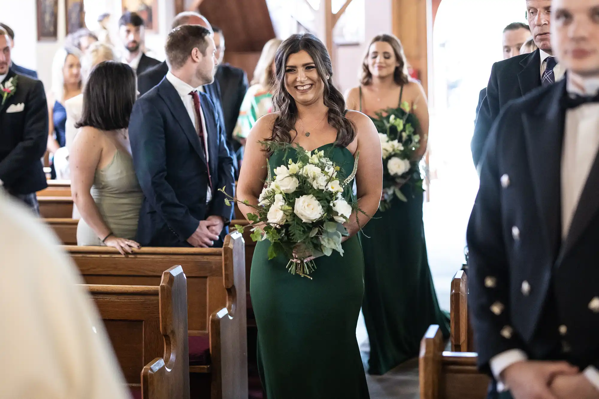 A bridesmaid in a green dress smiling as she walks down the aisle in a church, holding a bouquet, with guests looking on.