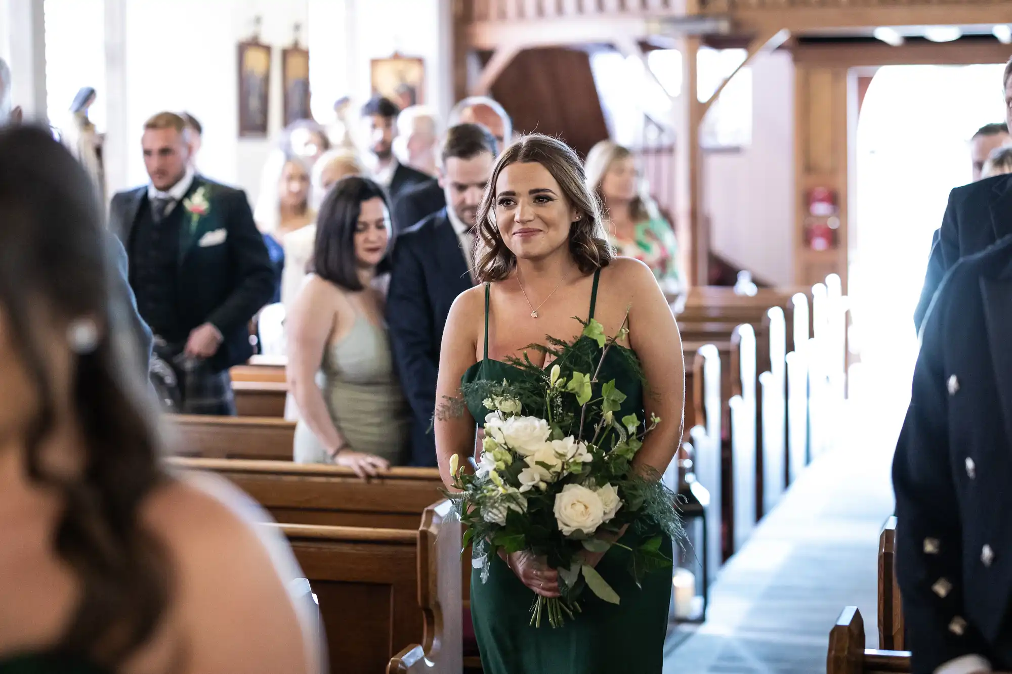 A bridesmaid in a green dress holding a bouquet walks down the aisle in a church, smiling, with guests watching.