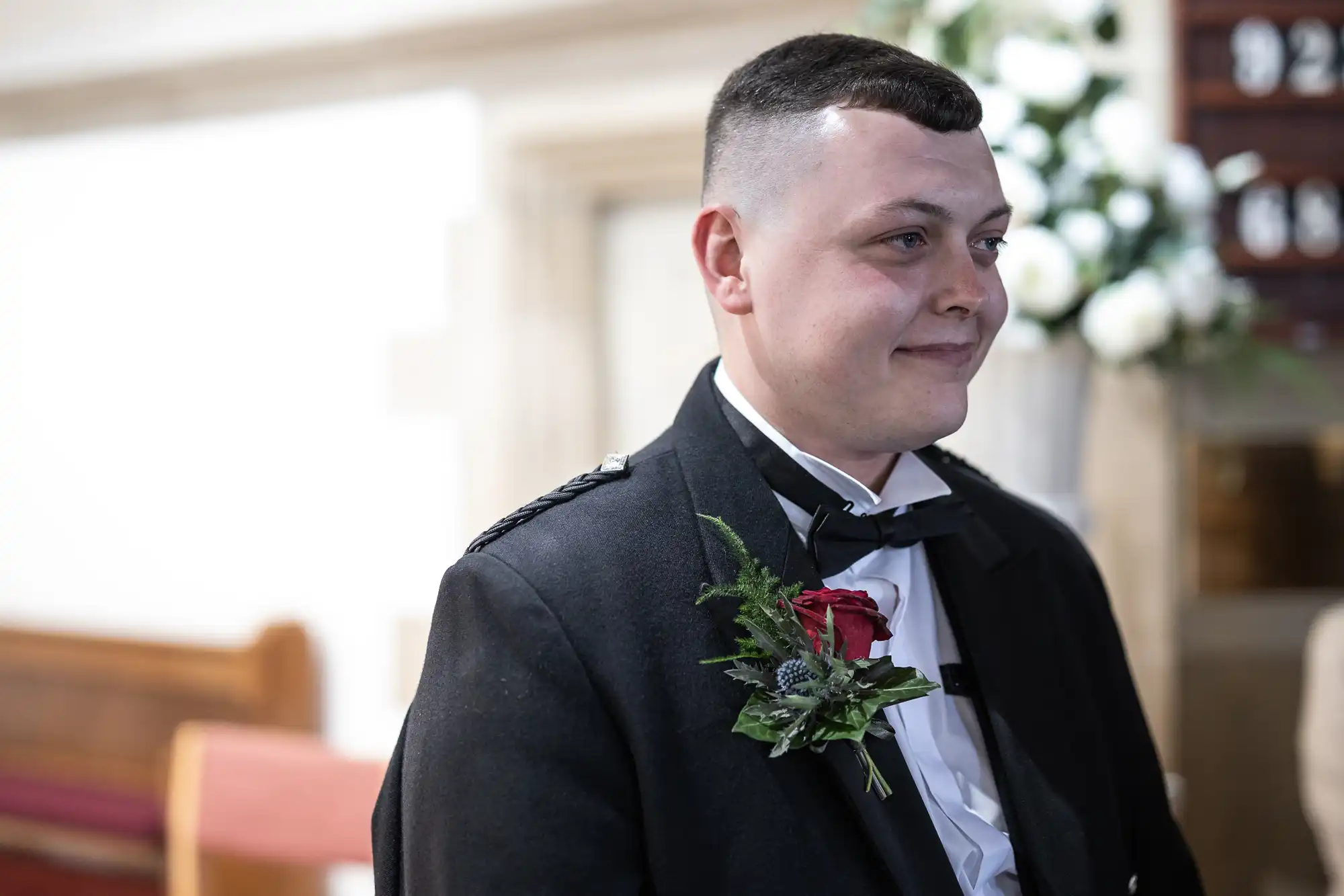 A man in a formal suit with a red rose boutonniere smiling inside a church.