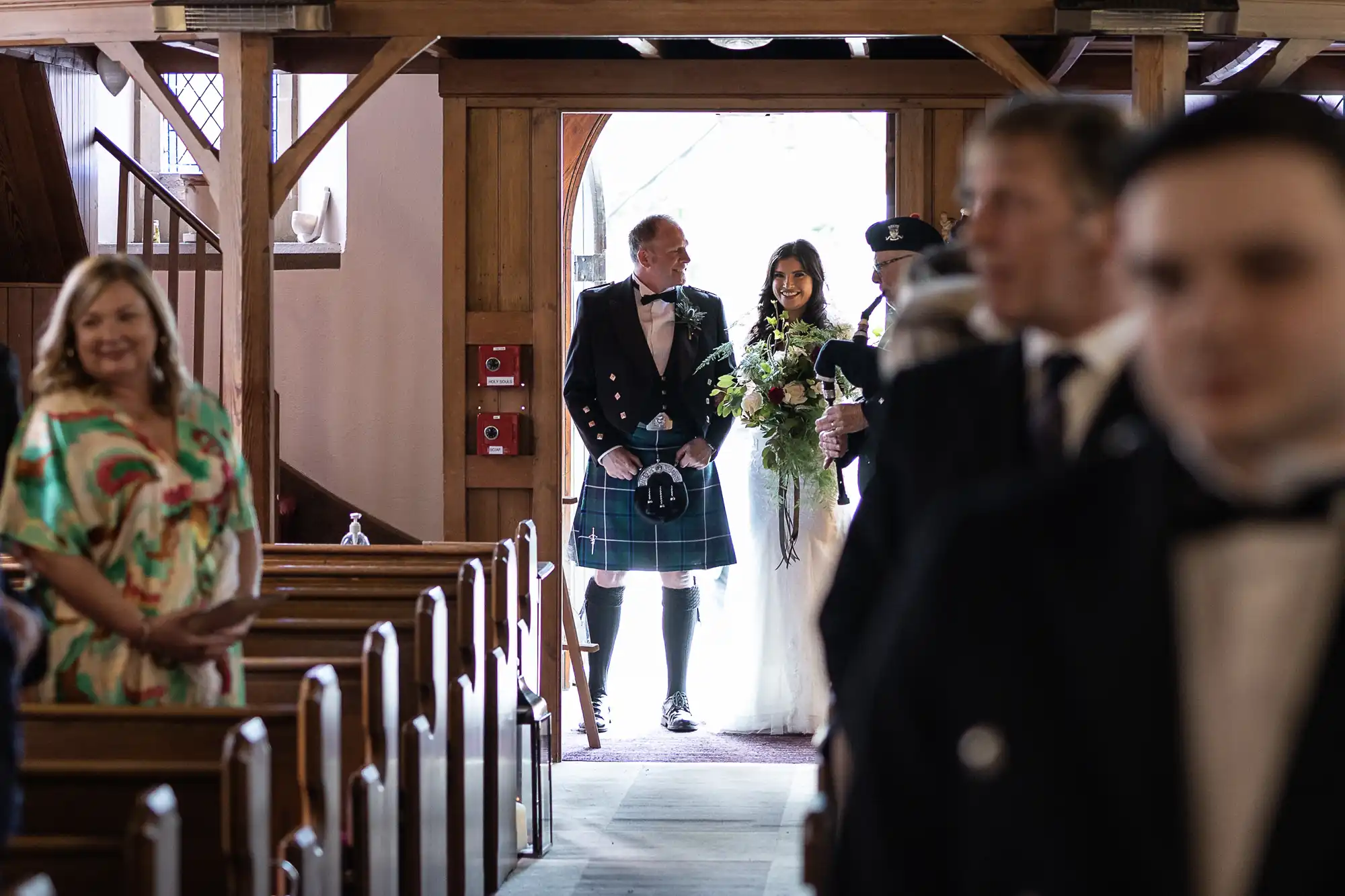 Bride and father in Scottish attire entering a church, viewed between rows of guests.