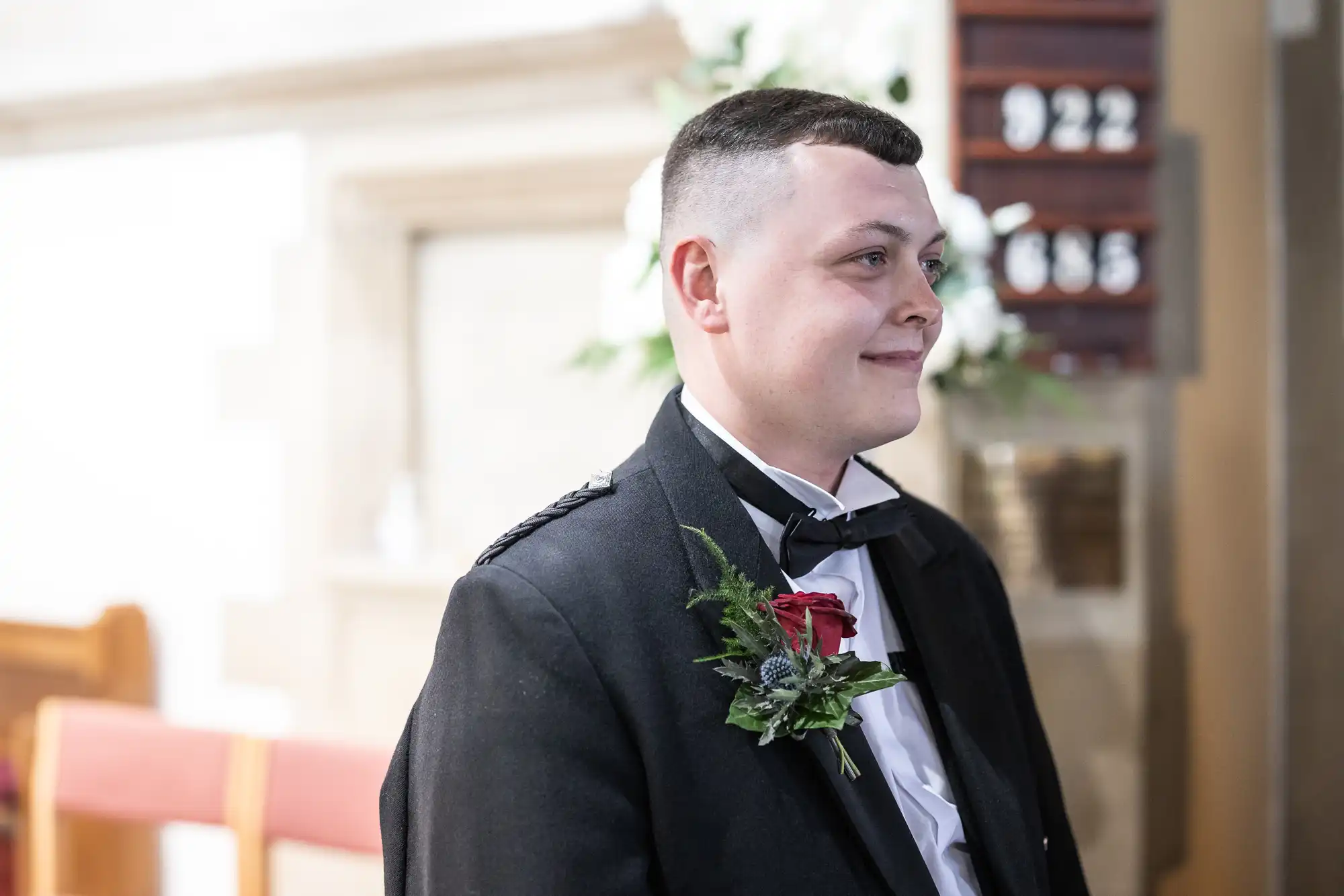 Groom in a black tuxedo with a red rose boutonniere smiling inside a church during a wedding ceremony.