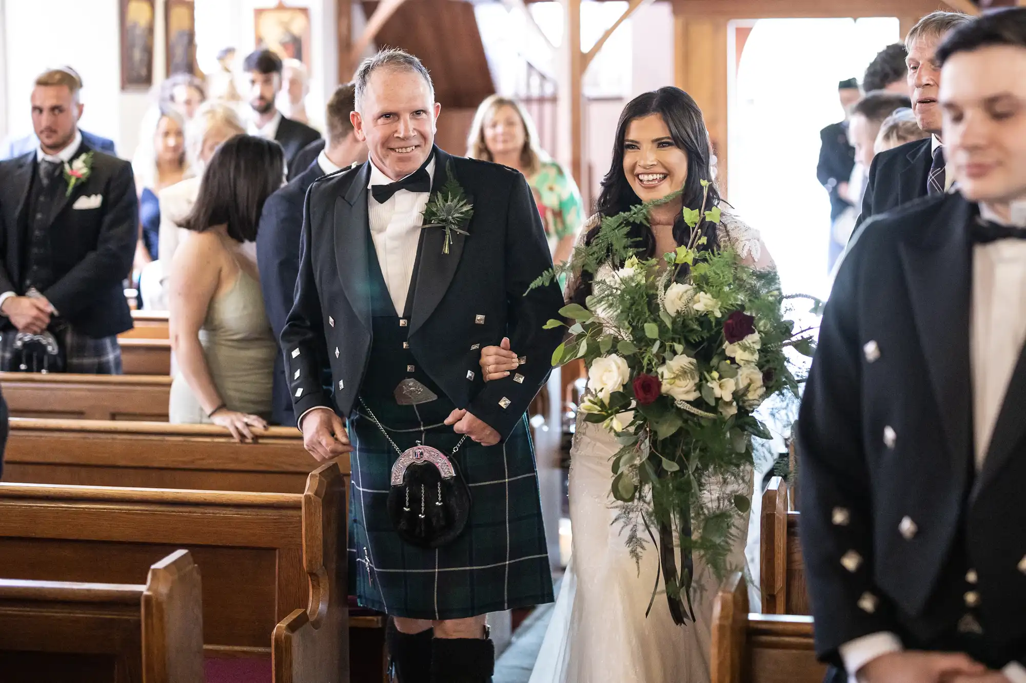 A bride and her father walking down the aisle, the father in a kilt and the bride holding a large bouquet, with guests watching from the pews.