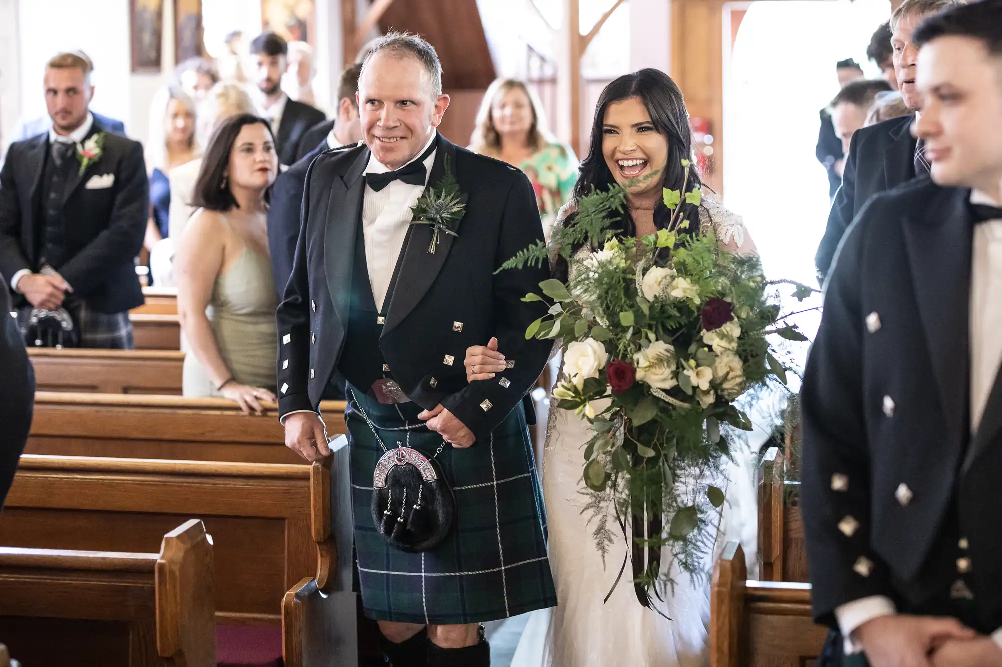 A bride and her father walking down the aisle in a church, both smiling, with the father wearing a traditional kilt and the bride holding a bouquet.