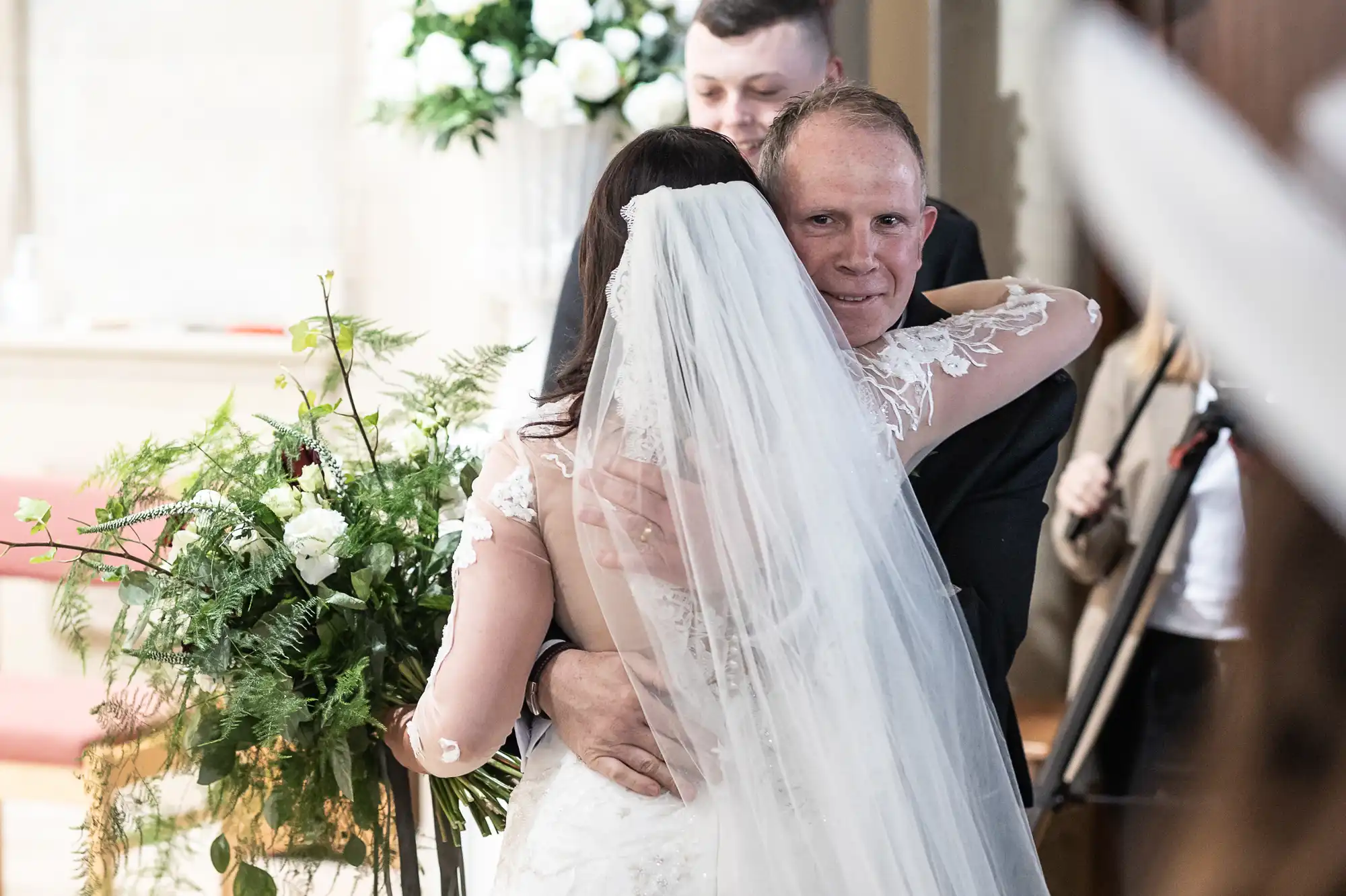 Father emotionally hugs a bride during a wedding ceremony in a church, surrounded by floral decorations.