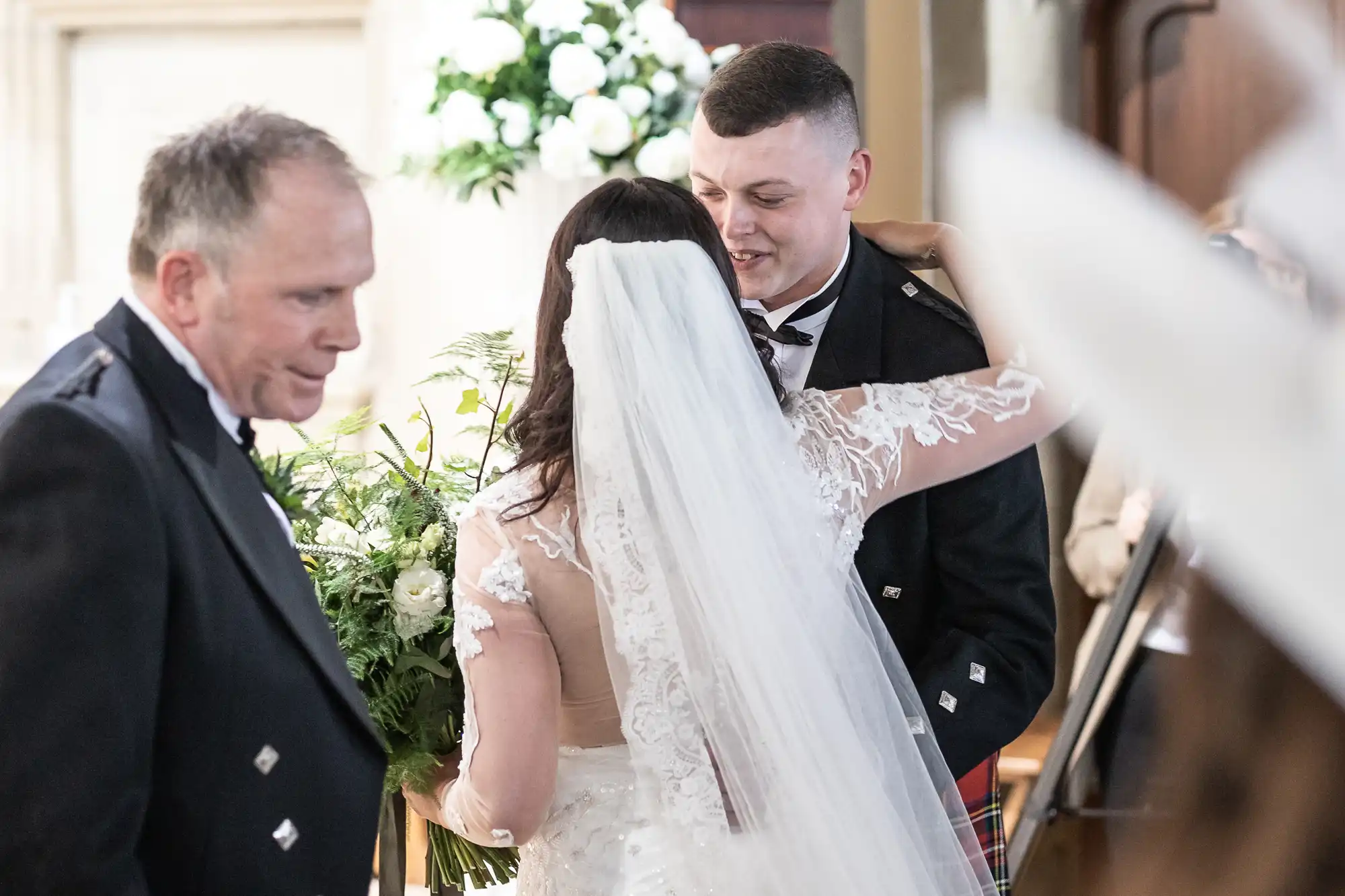 A bride and groom embrace, smiling, as an older man looks on during a wedding ceremony in a church.