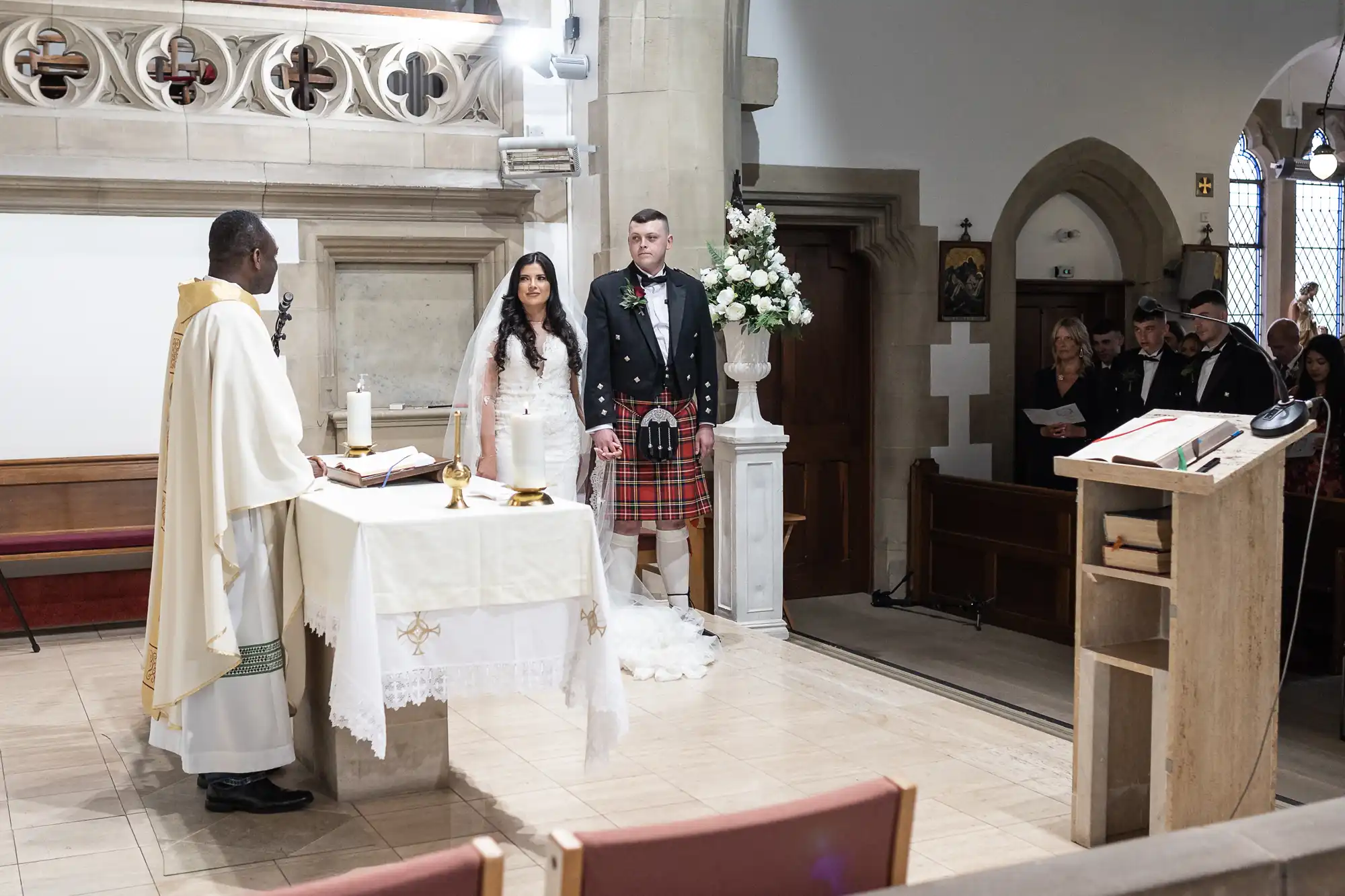 A bride and groom stand before a priest at a church altar during a wedding ceremony; the groom wears a kilt. Guests are seated in the background.