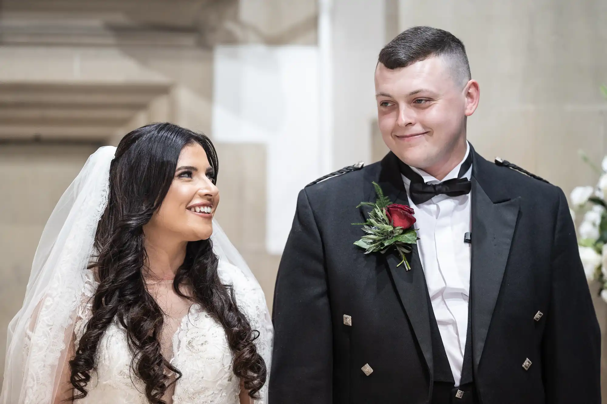 A joyful bride in a white lace gown and veil smiles at someone off-camera, while a groom in a black military uniform with a boutonniere looks at her fondly.