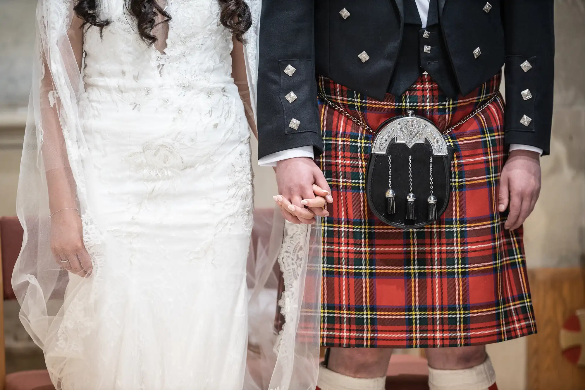 A bride in a white dress and a groom in a tartan kilt holding hands at a wedding ceremony.