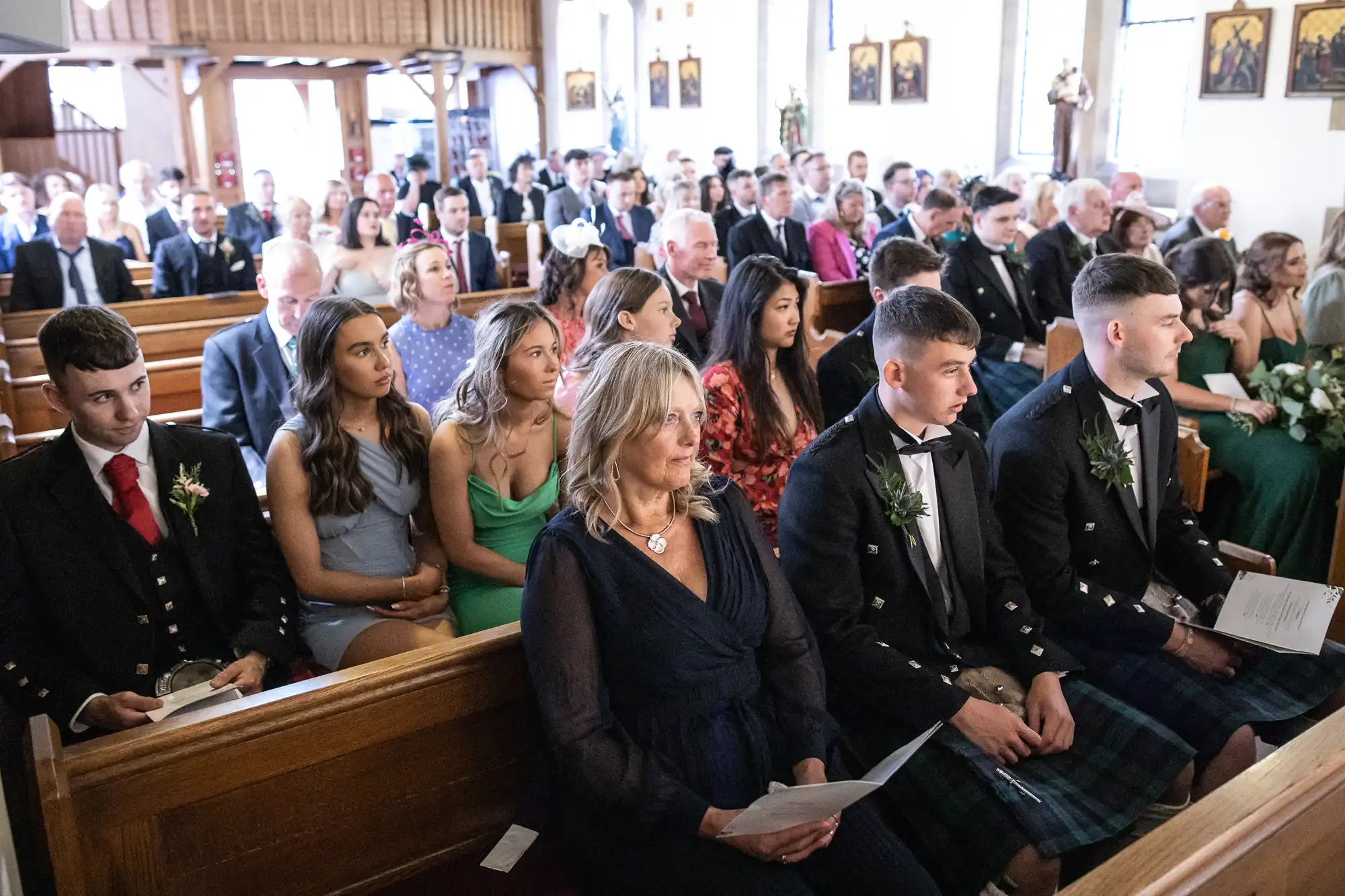 A congregation of formally dressed people attending a church ceremony, some in suits and one in a traditional kilt.