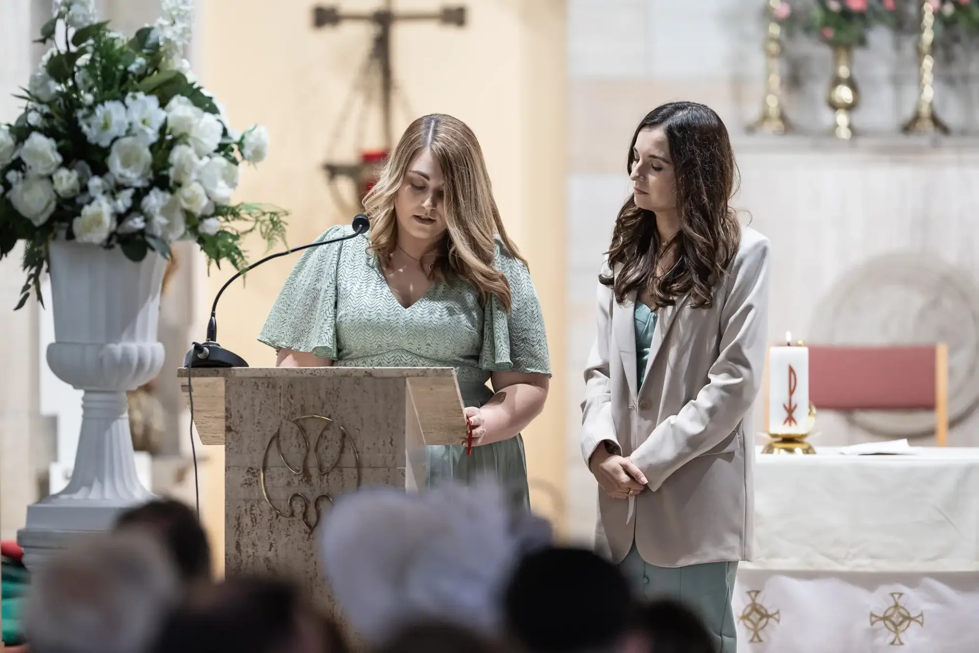 Two women speaking at a podium in a church, with one of them reading from a notebook, as attendees listen in the background.