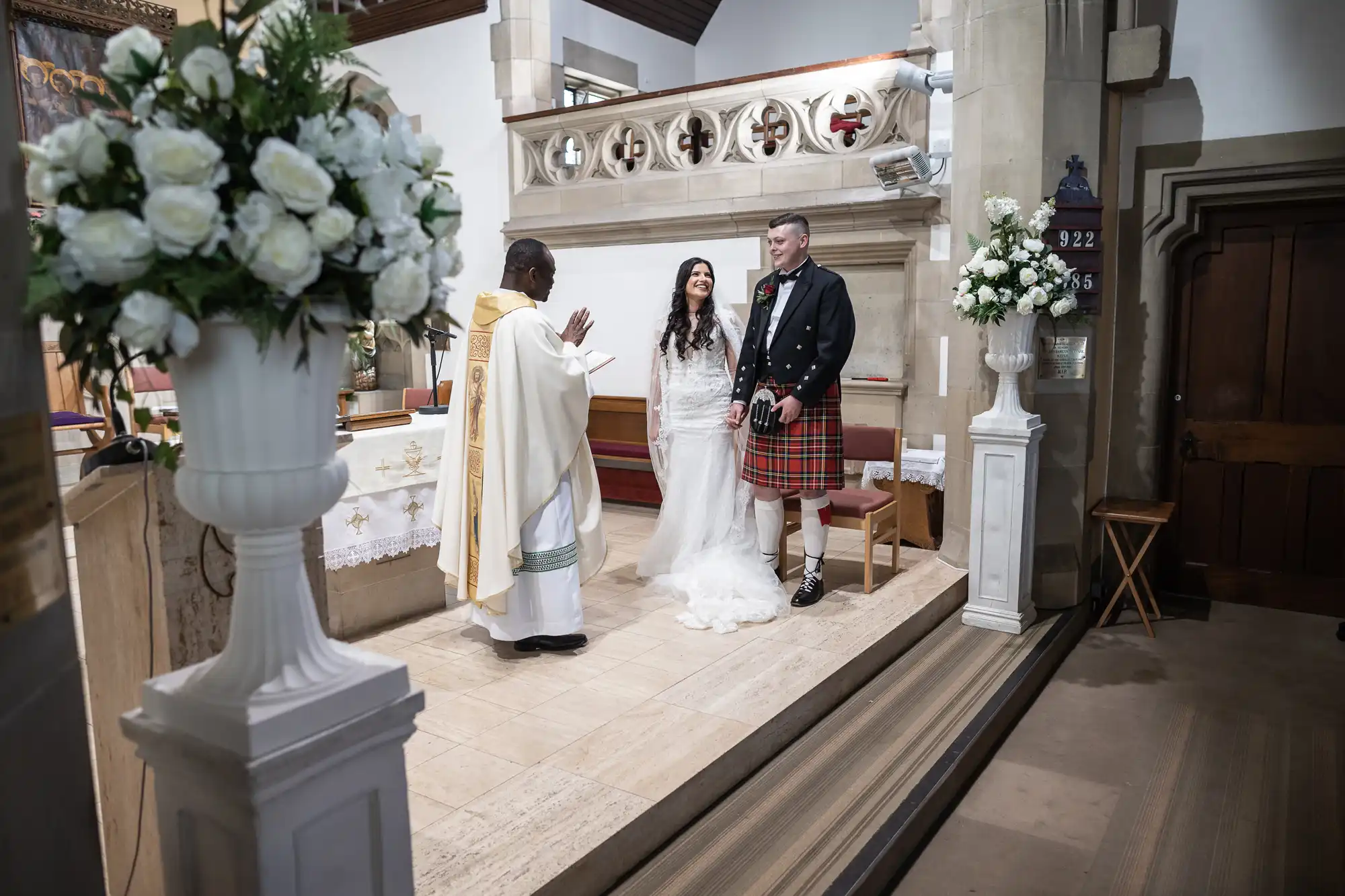 A bride and groom stand before a priest inside a church during a wedding ceremony; the groom wears a kilt, and floral decorations are visible.