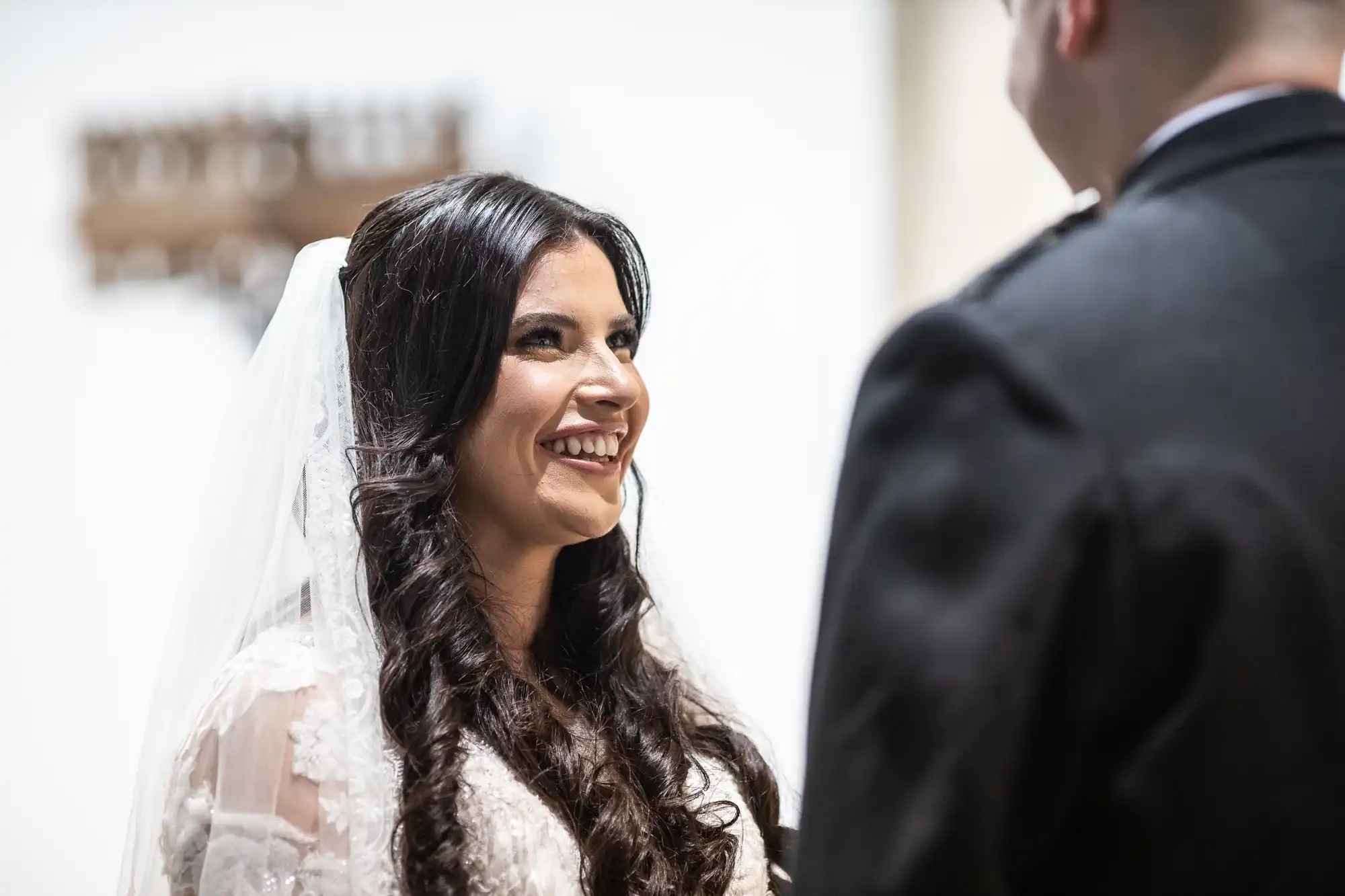 A bride in a white lace gown and veil smiles joyfully at her groom in a formal setting.