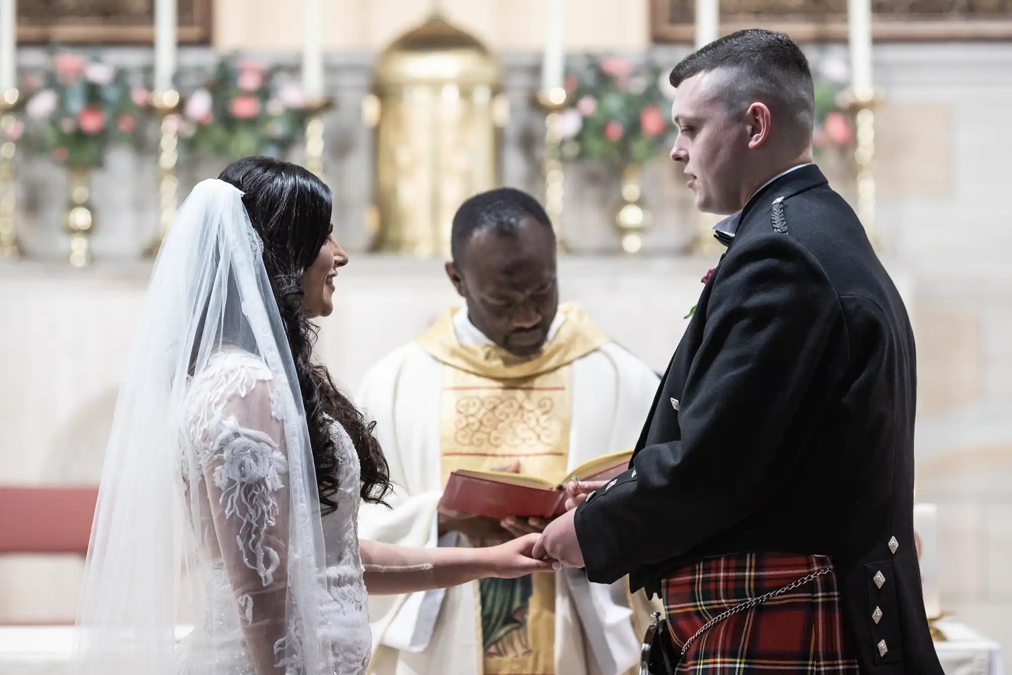 A bride and groom in wedding attire, with the groom wearing a kilt, exchange vows before a priest in a festively decorated church.
