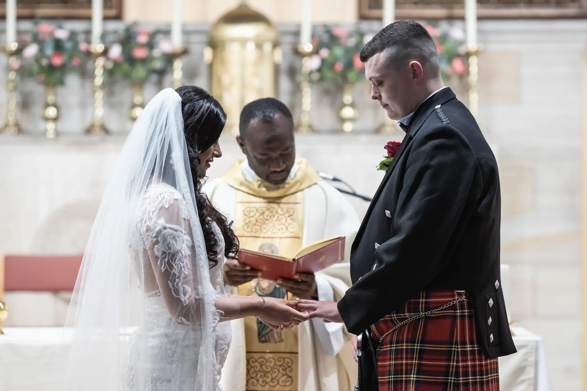 A bride in a white gown and a groom in a kilt holding hands during a wedding ceremony officiated by a priest in a church.