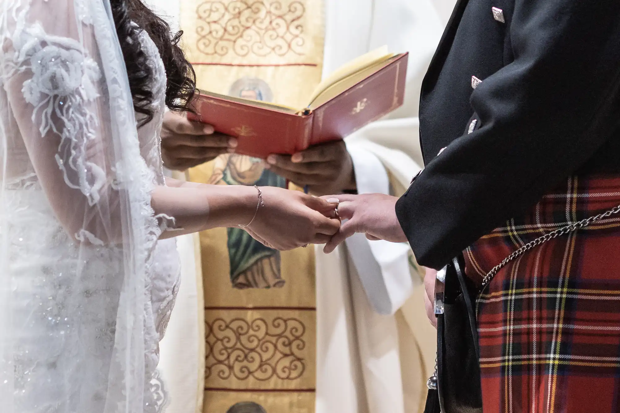 A bride in a white dress and a groom in a kilt exchanging rings during a wedding ceremony, with a clergy member holding a red book.