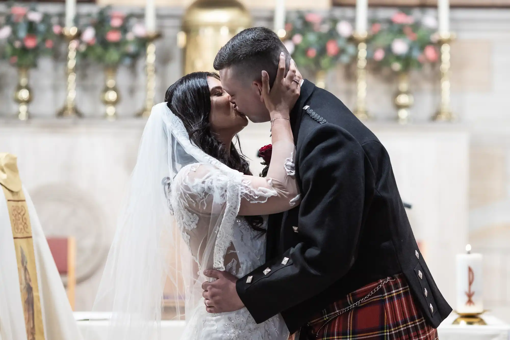 A bride in a white dress and a groom in a kilt passionately kiss in a church decorated with flowers.