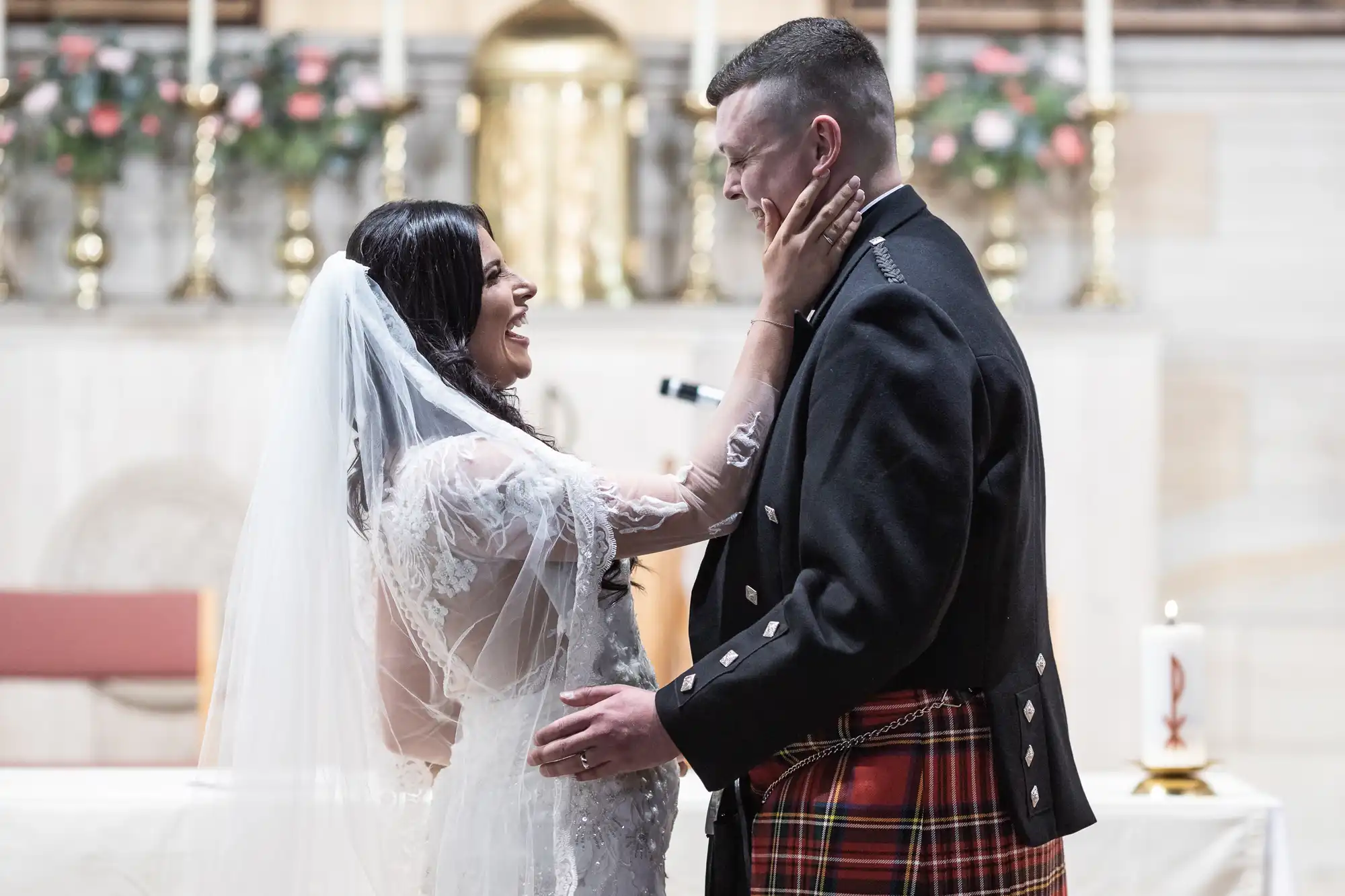 A bride in a white gown and veil tenderly touches the face of her groom in a traditional kilt inside a church.