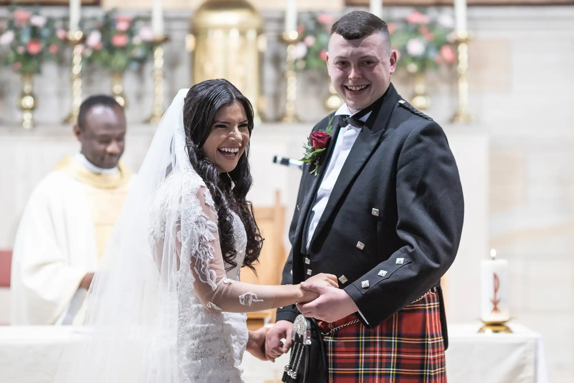 A joyful bride and groom exchange rings in a church wedding ceremony, with the groom wearing a kilt and the bride in a white lace dress. A priest observes in the background.