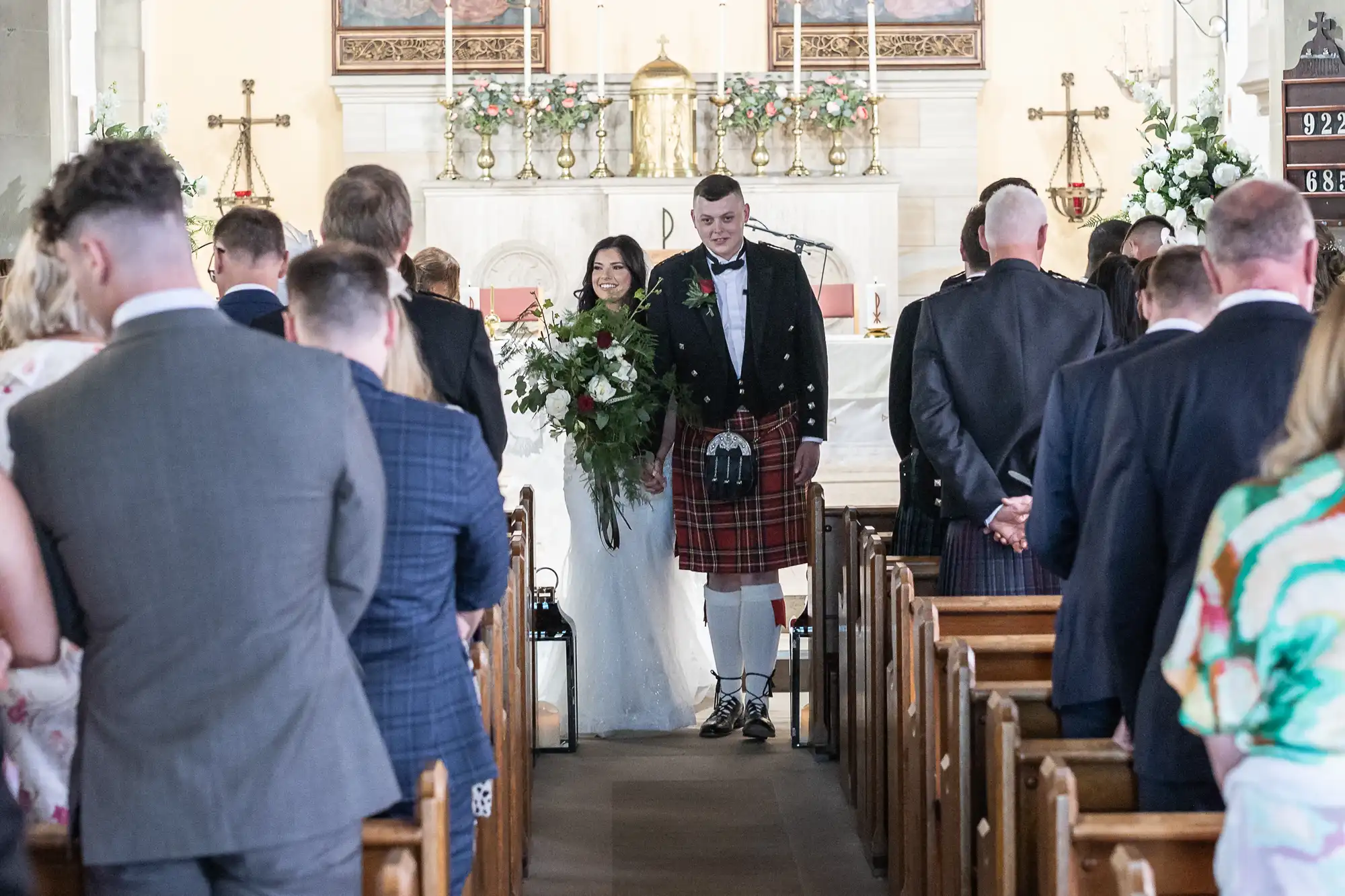 Bride and groom smiling as they walk down the aisle in a church, surrounded by guests; groom is wearing a traditional kilt.