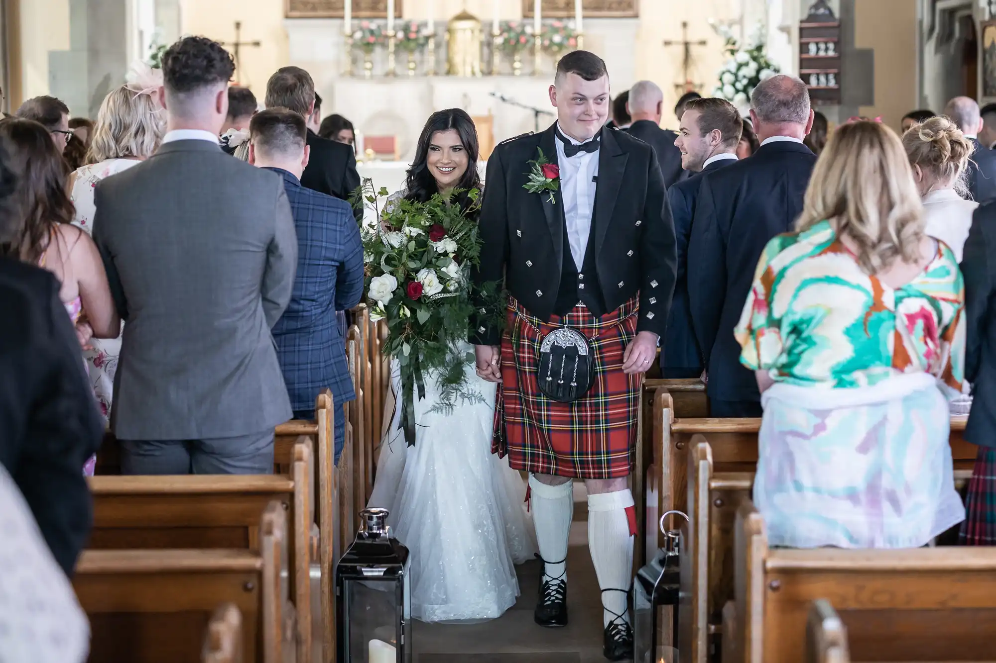 Bride and groom walking down the aisle, the groom in a kilt, surrounded by guests in a church.