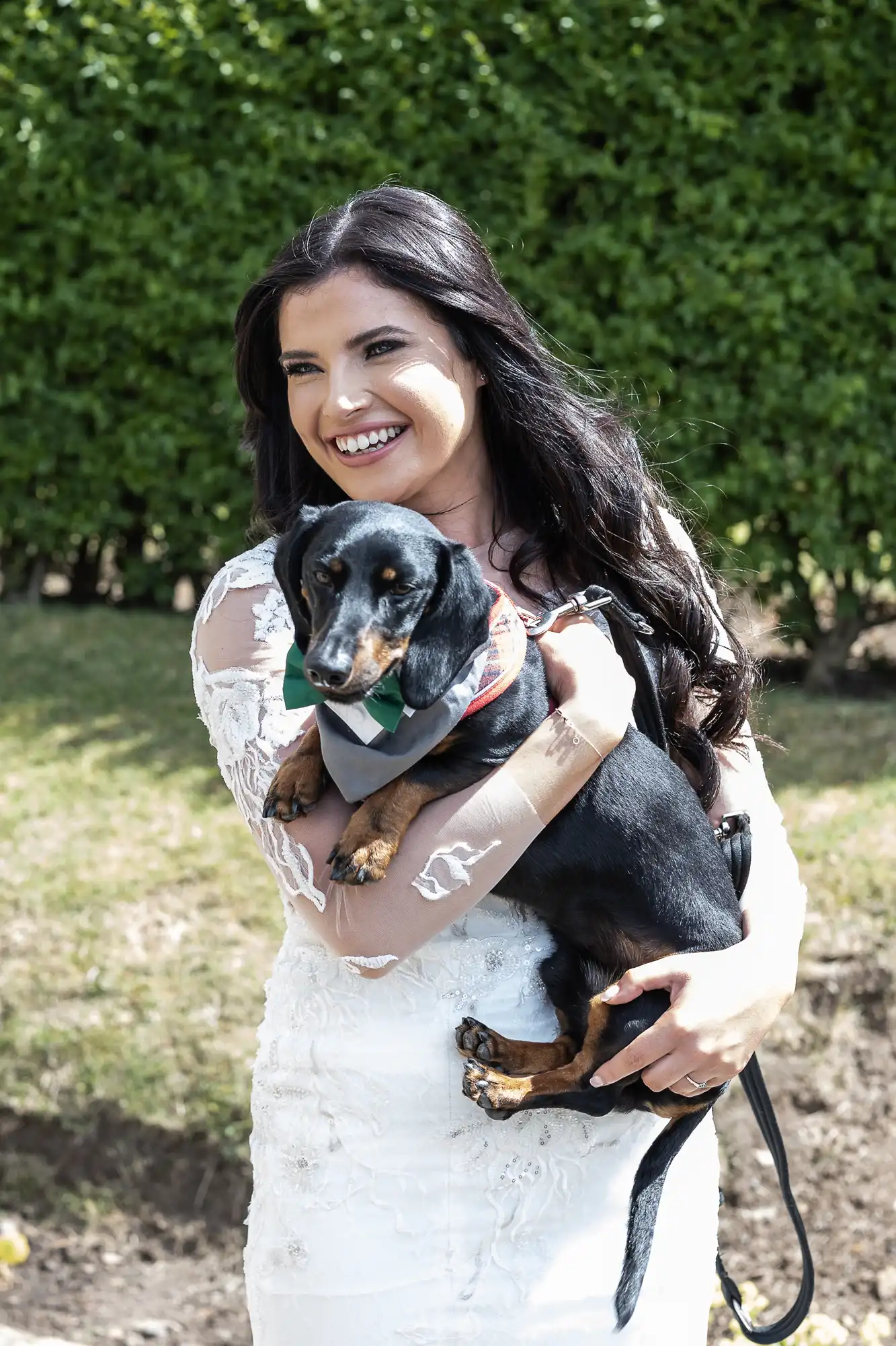 A woman in a white dress smiles broadly while holding a dachshund in a harness at a sunny outdoor setting.