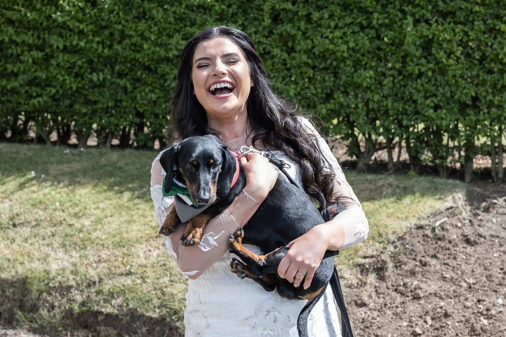 Woman in a white dress laughing joyfully while holding a black dachshund in a sunny garden setting.
