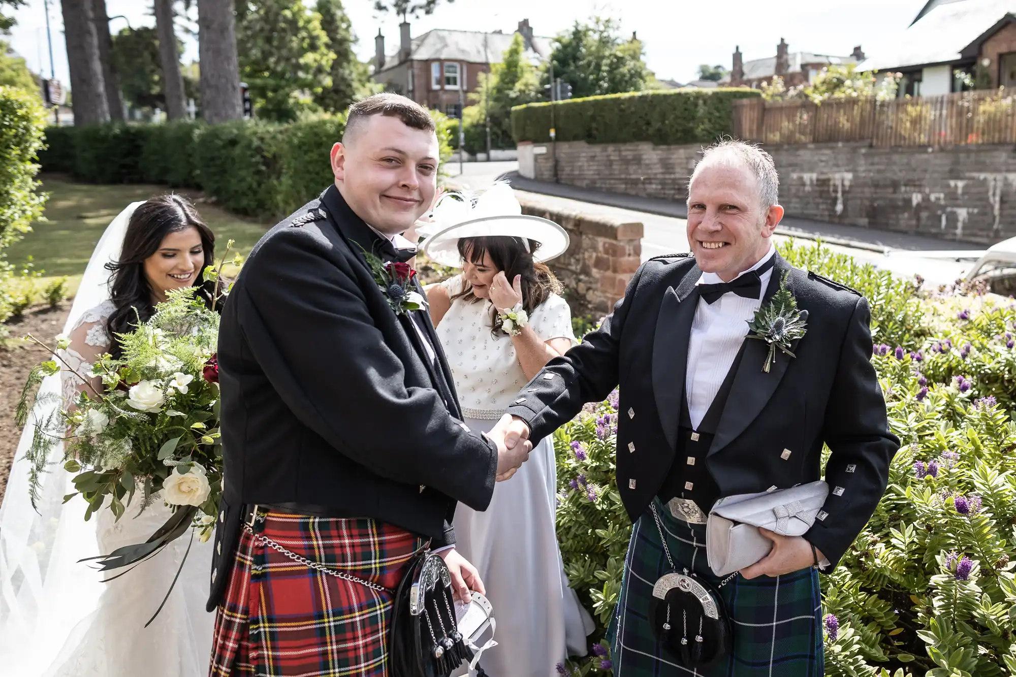 A groom in a kilt shaking hands with father of the bride in a kilt at a wedding, with a bride and another woman in the background.