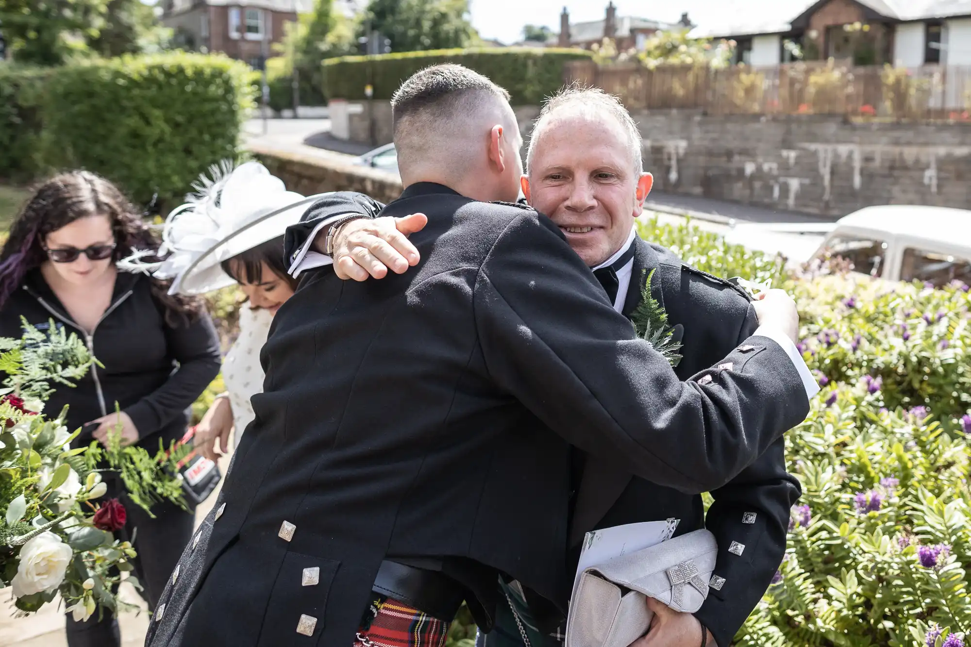 Two men embracing warmly at an outdoor gathering, one wearing a kilt and the other in a suit, with people and floral decorations in the background.