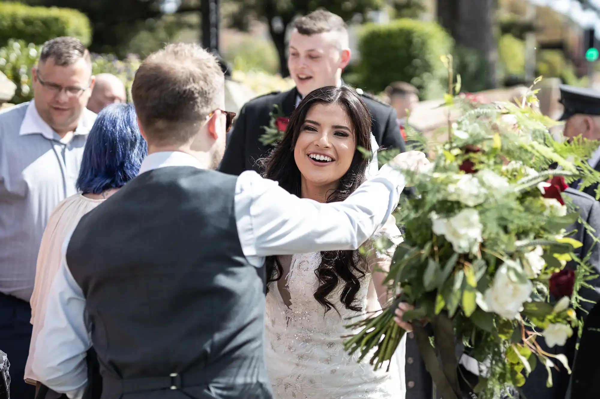A bride in a white dress smiling and reaching out to a groom, surrounded by guests and floral arrangements on a sunny day.
