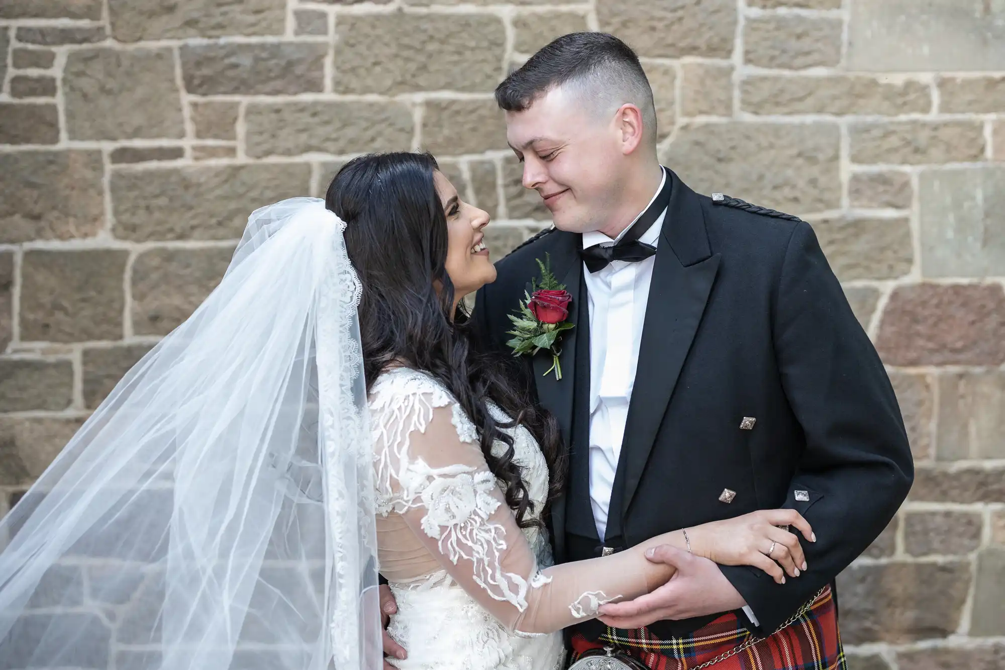 Bride in a white lace gown and groom in a kilt smiling at each other affectionately, standing against a stone wall backdrop.