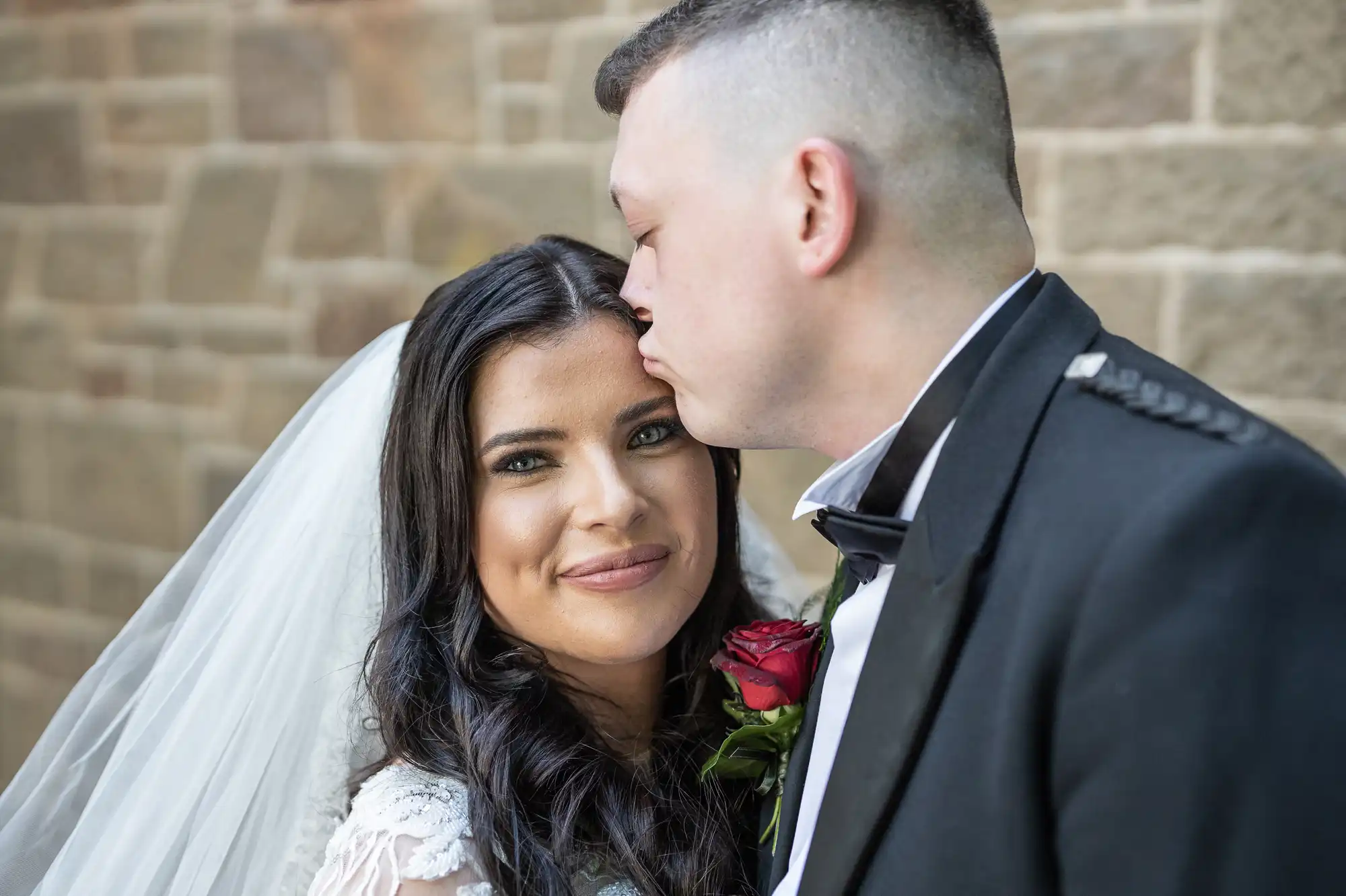 Groom in a military uniform kissing bride on the forehead outside a stone building, bride smiling at camera.