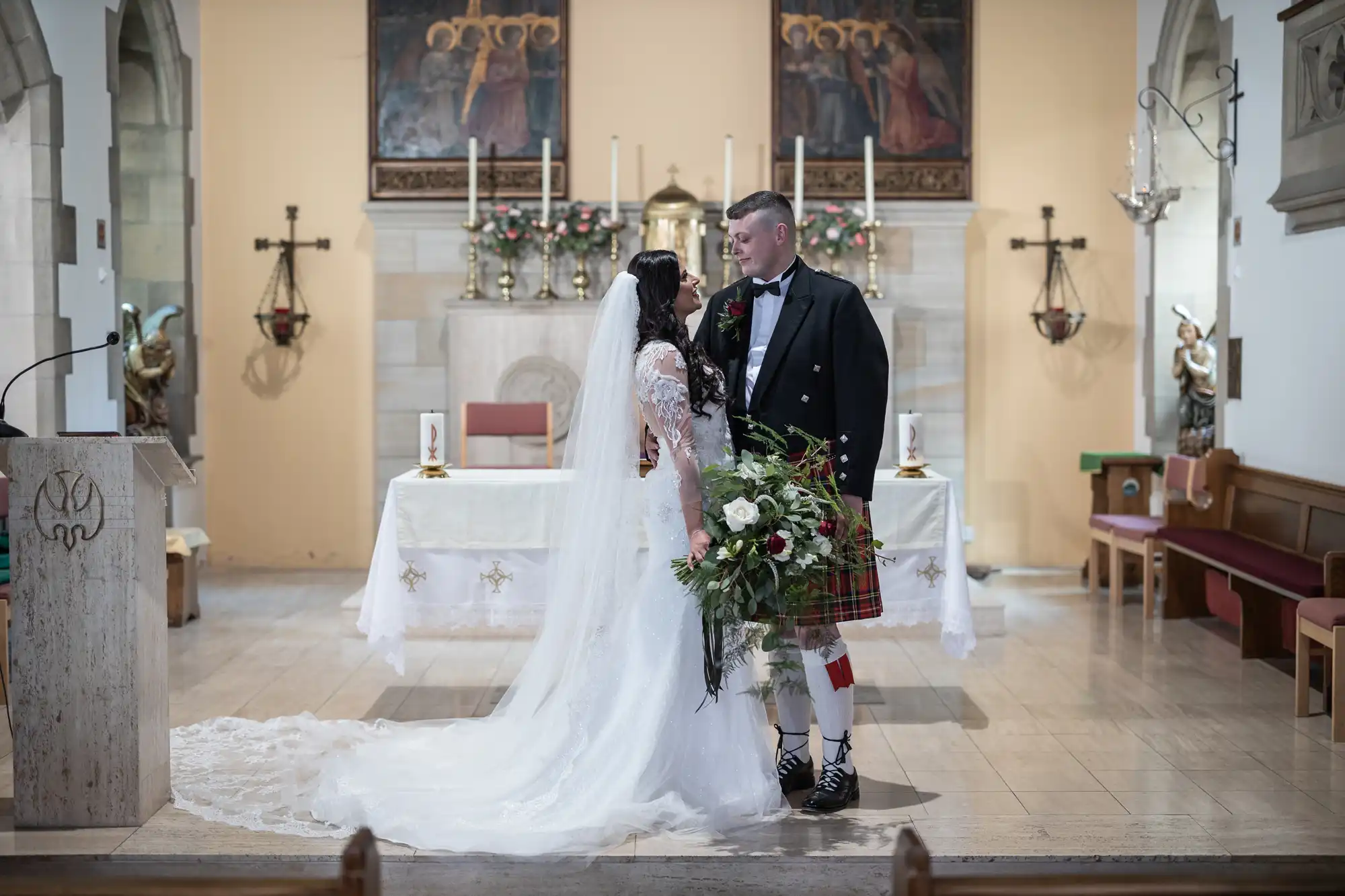 A bride in a long white dress and a groom in a kilt holding hands in a church, smiling at each other lovingly.
