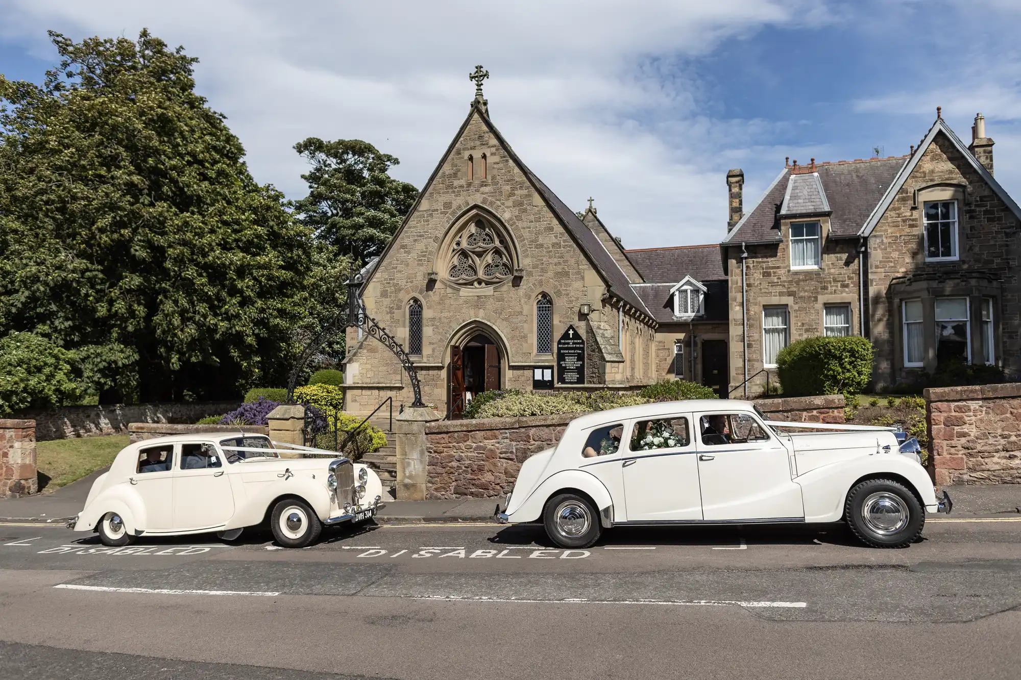 Two vintage white cars parked in front of a quaint stone church with a "Disabled" parking sign on the road.