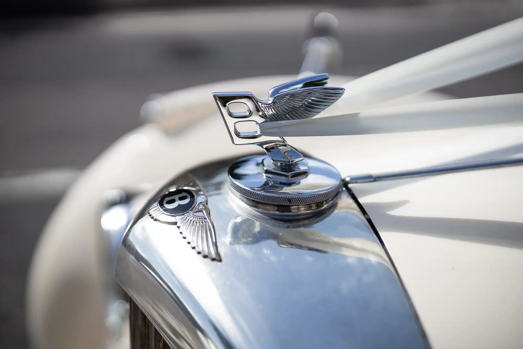 Close-up of a Bentley car's hood ornament and logo, featuring a pair of wings and the letter B, mounted on a chrome grille of a vintage car.