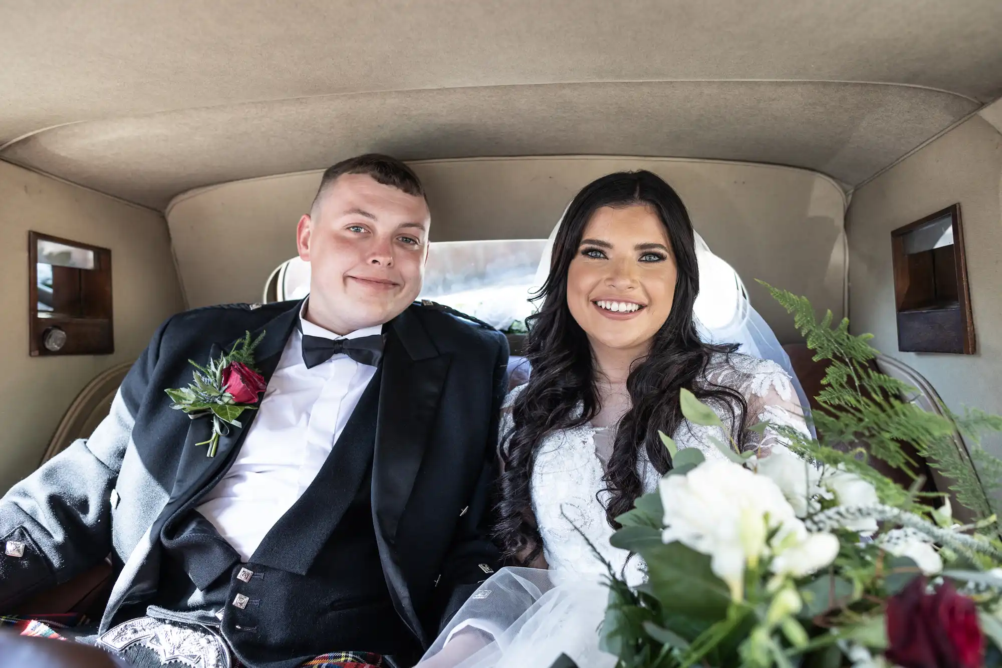 A bride and groom smiling inside a vintage car, with the bride holding a bouquet of flowers.