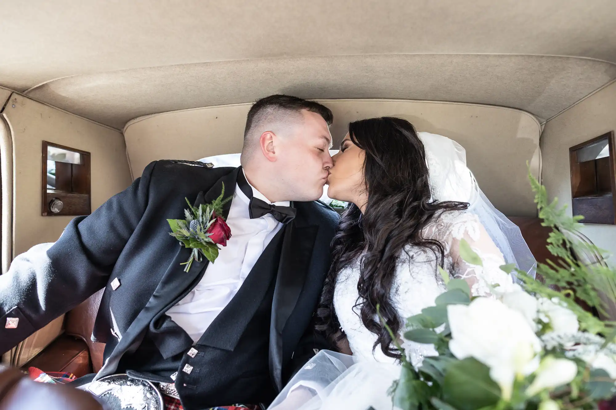 A newlywed couple kissing inside a vintage car, surrounded by white flowers, with the bride in a white dress and the groom in a black tuxedo.