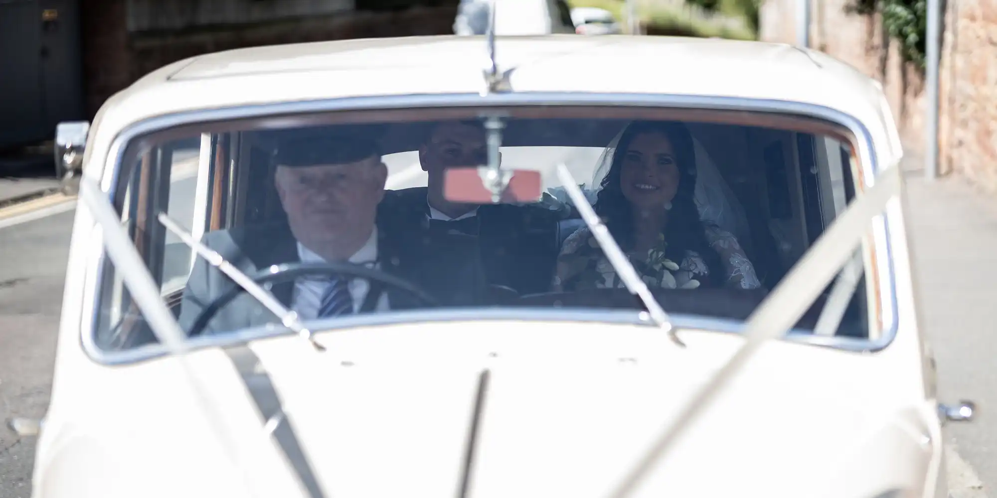 A bride and a chauffeur seen through the windshield of a classic car, with a focus on the bride smiling.