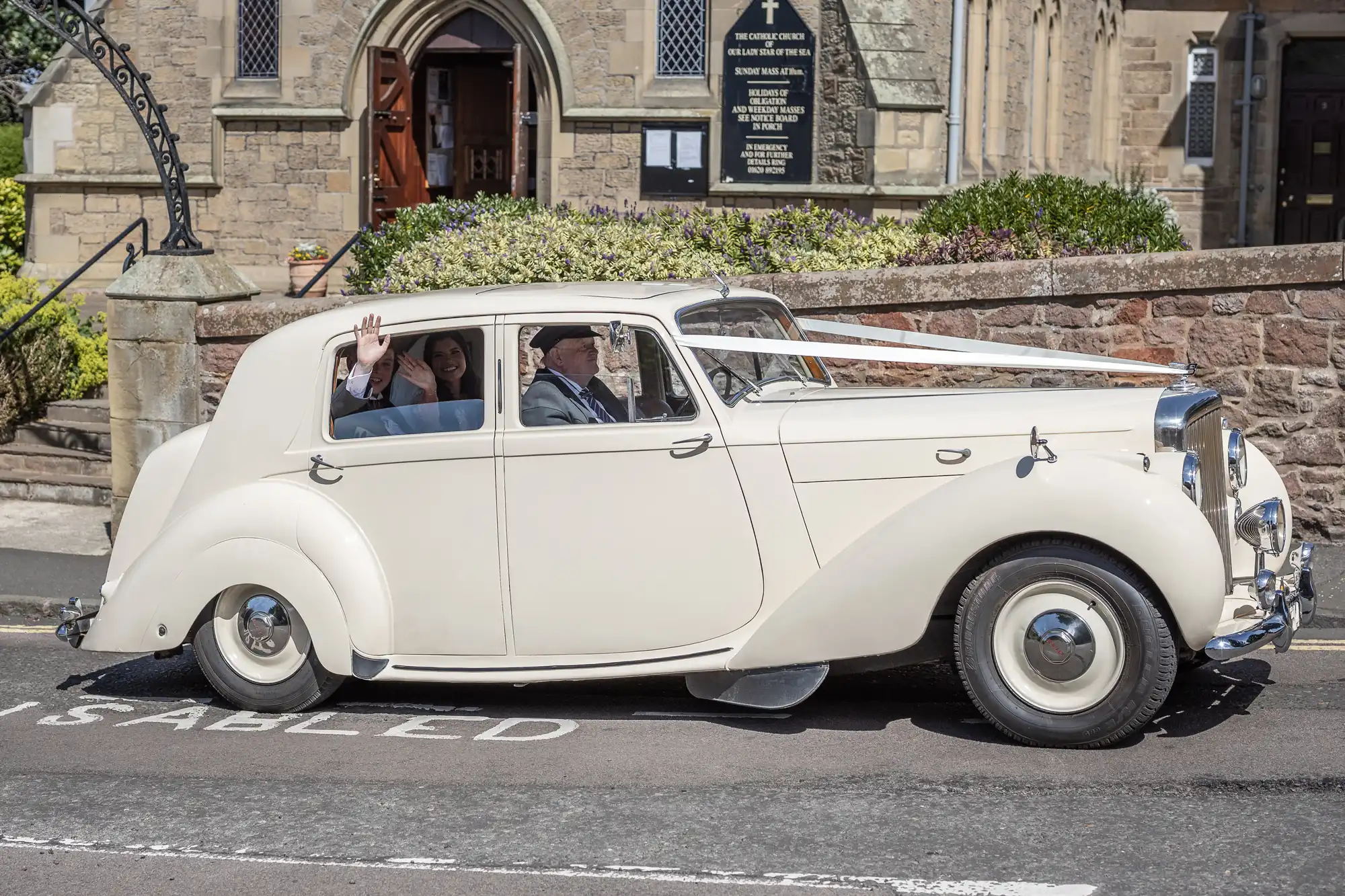 A vintage white wedding car driving down the street with a couple waving from the back seat.