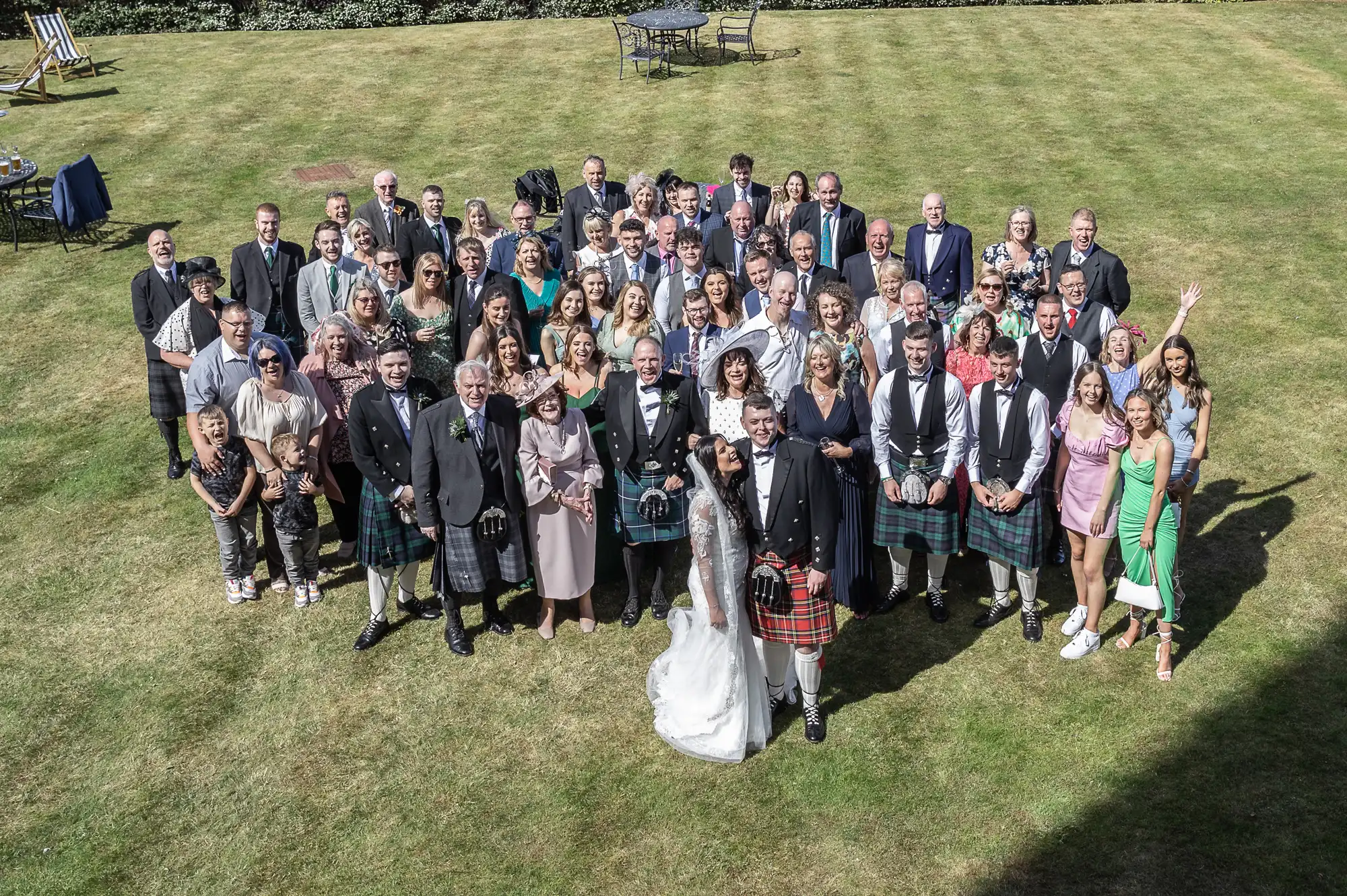 A large wedding group, including guests in suits and kilts, posing outdoors on a lawn with the bride and groom at the center.