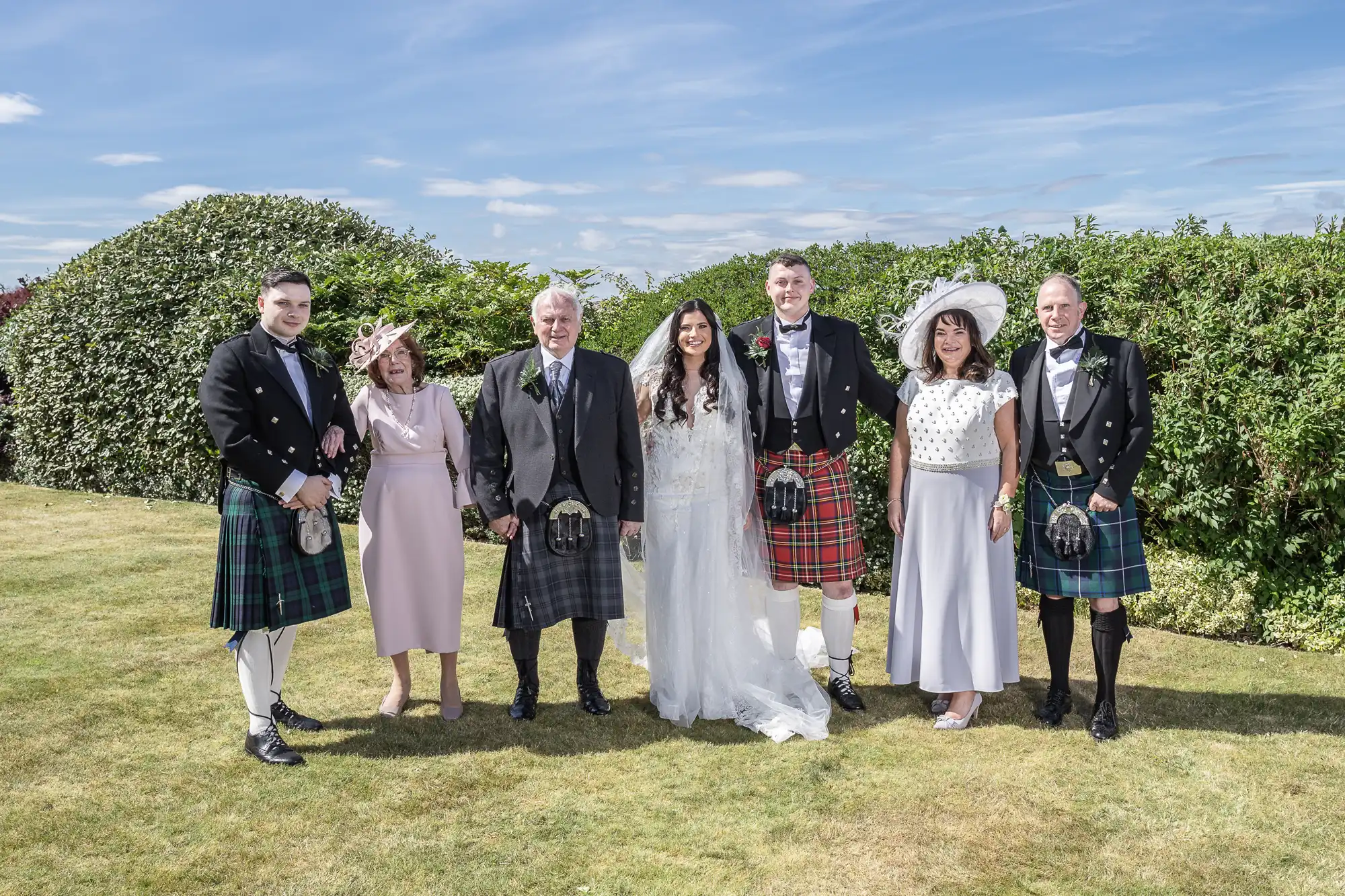 Bride and groom with four guests wearing traditional Scottish attire, posing on a lawn under a clear sky.