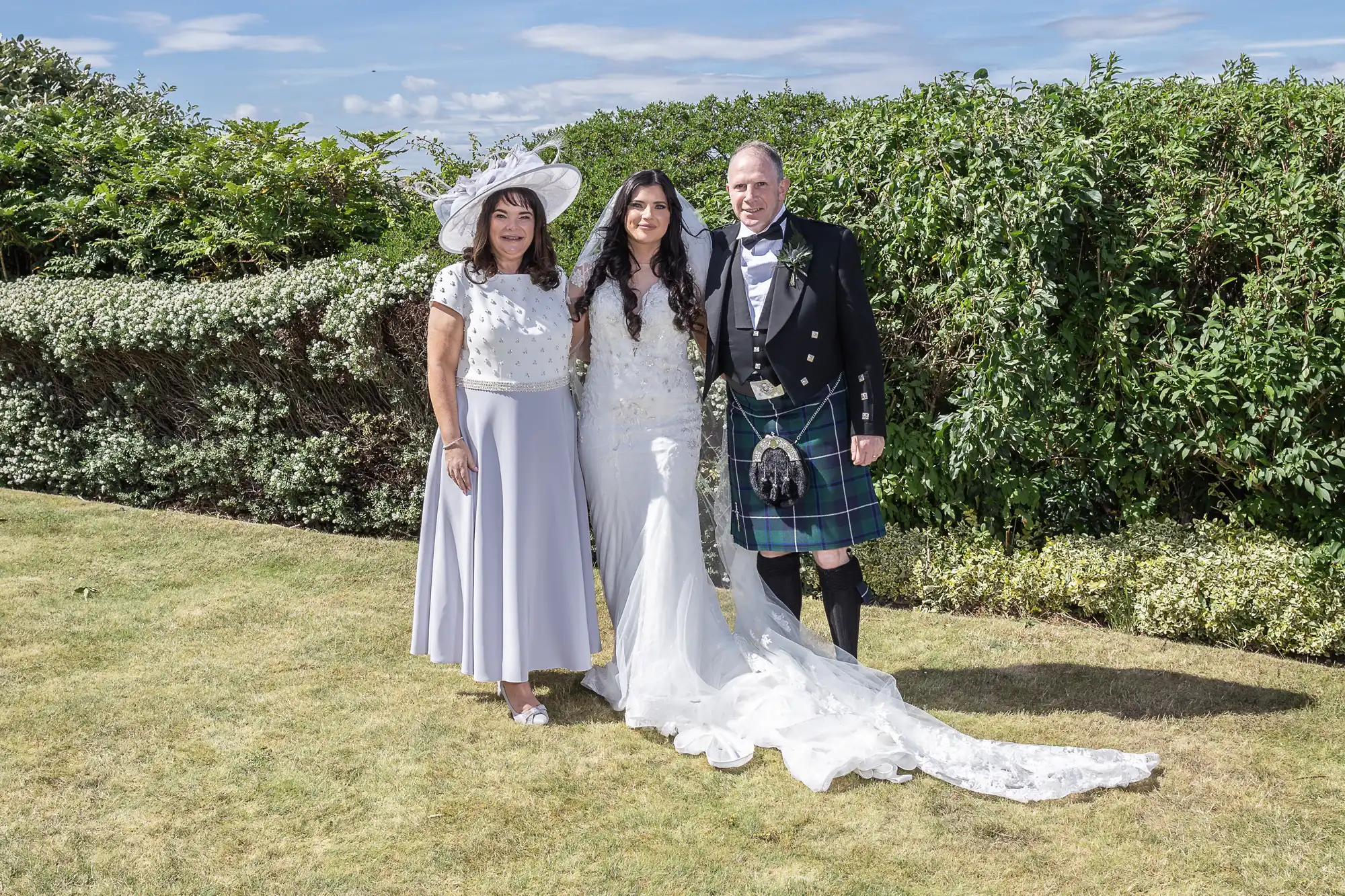 A bride in a white gown with a train stands next to a man in a kilt and an older woman in a white midi dress and hat, all smiling in front of a hedge under a clear sky.