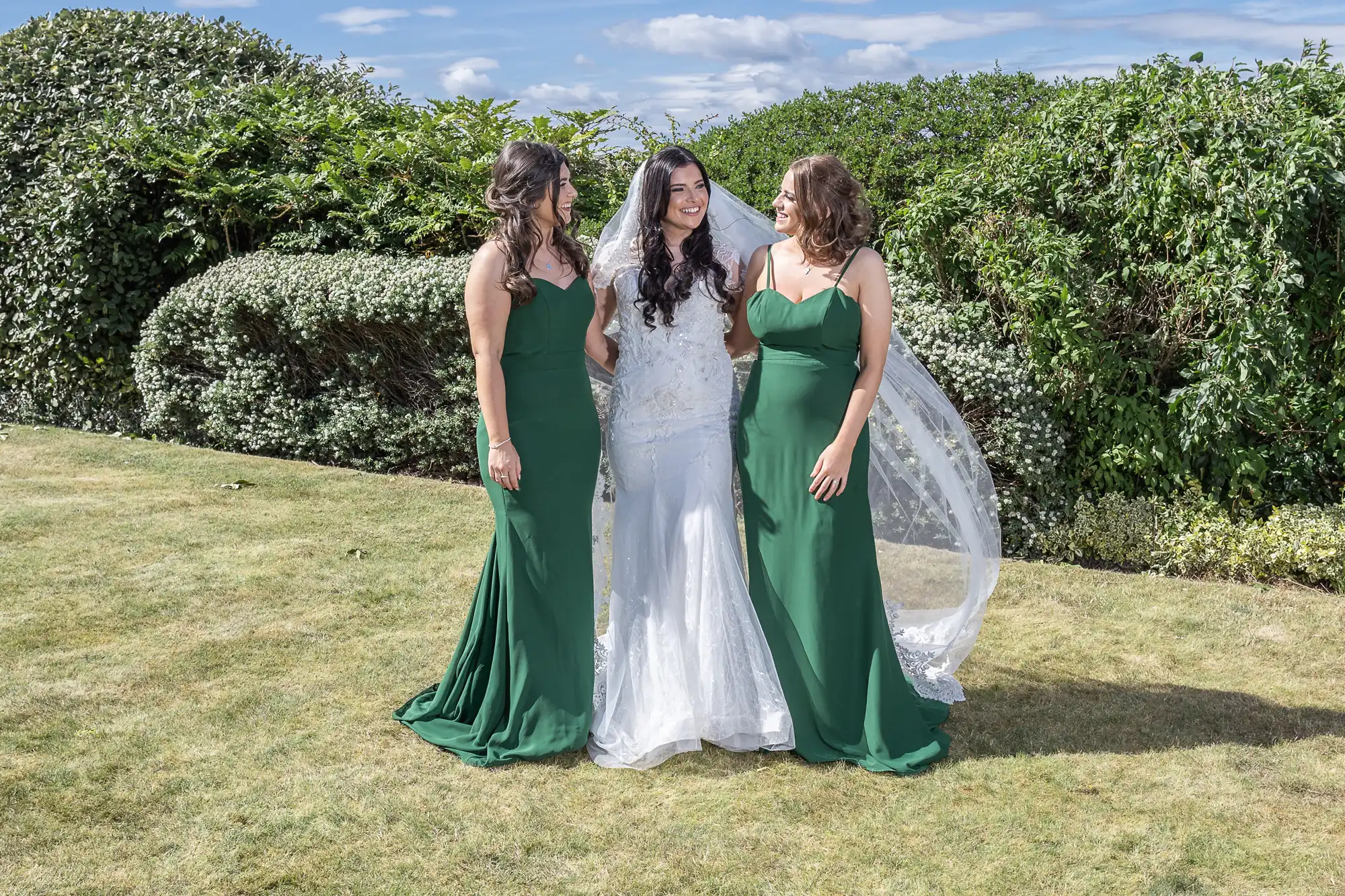 Three women dressed in formal attire, with the bride in white and two bridesmaids in green, standing and smiling on a lawn with a hedge in the background.