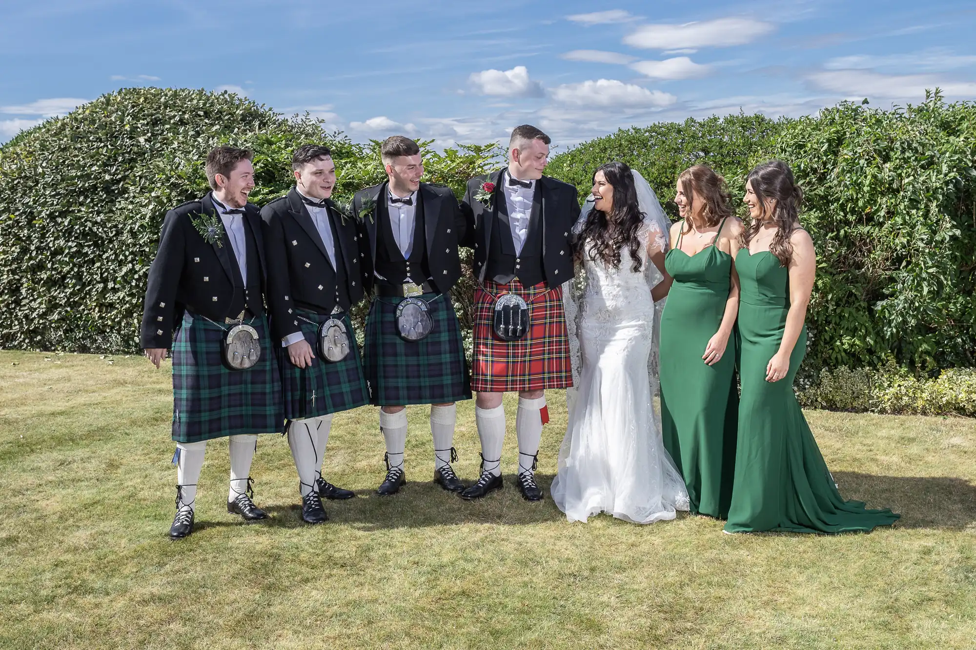 A bride in a white dress and groom in a tartan kilt with four groomsmen and two bridesmaids, all smiling in a lush garden.