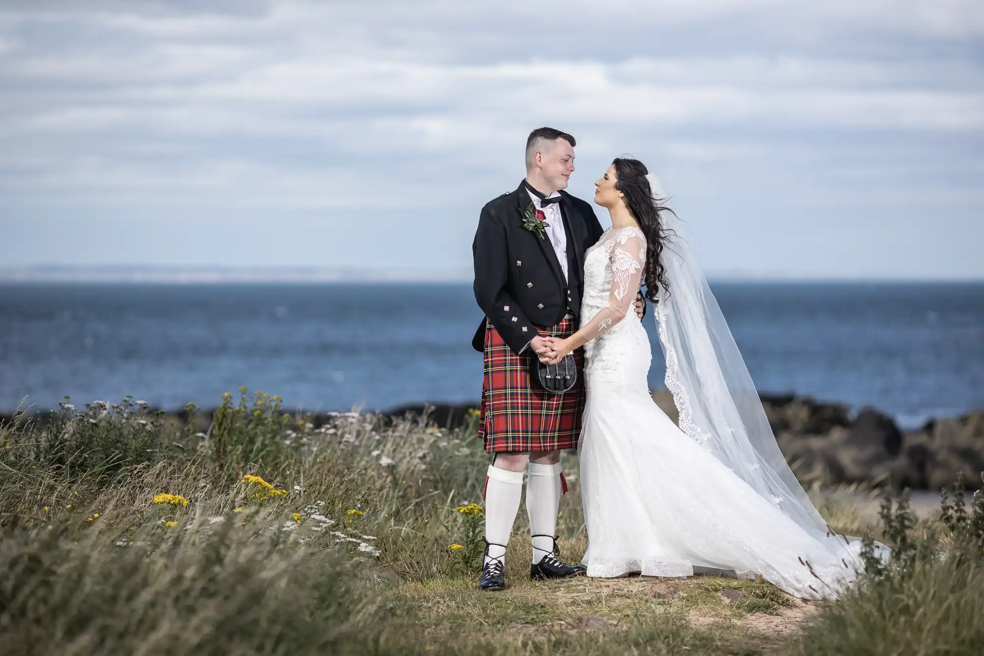 A bride and groom in wedding attire share a tender moment by the seaside, with the groom wearing a traditional kilt. Marine Hotel wedding photographers.
