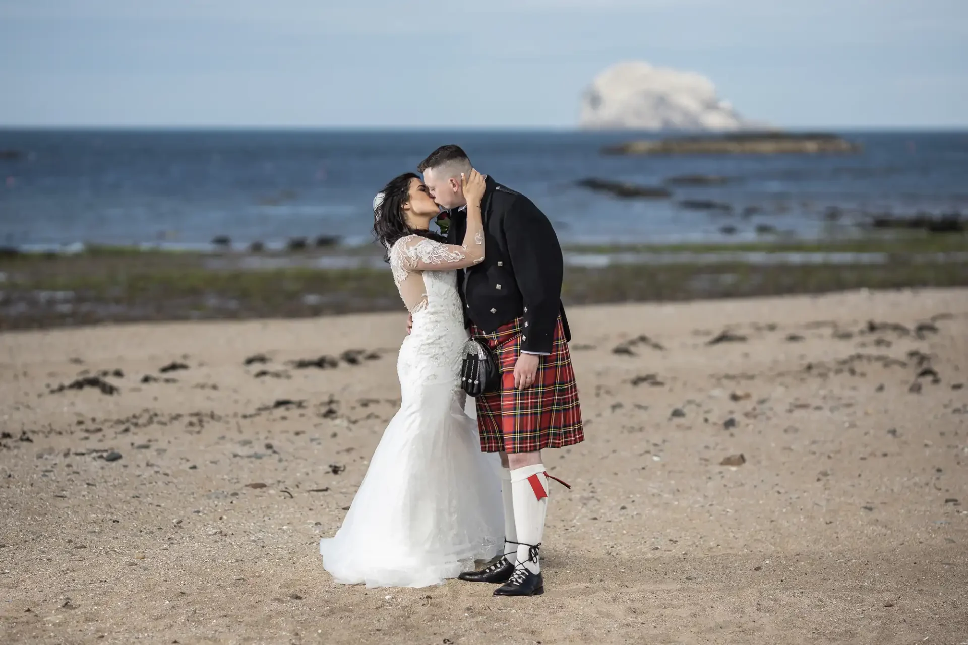A bride and groom kissing on a beach during their Marine Hotel wedding, with the groom wearing a kilt and the ocean and a rocky Bass Rock island in the background.