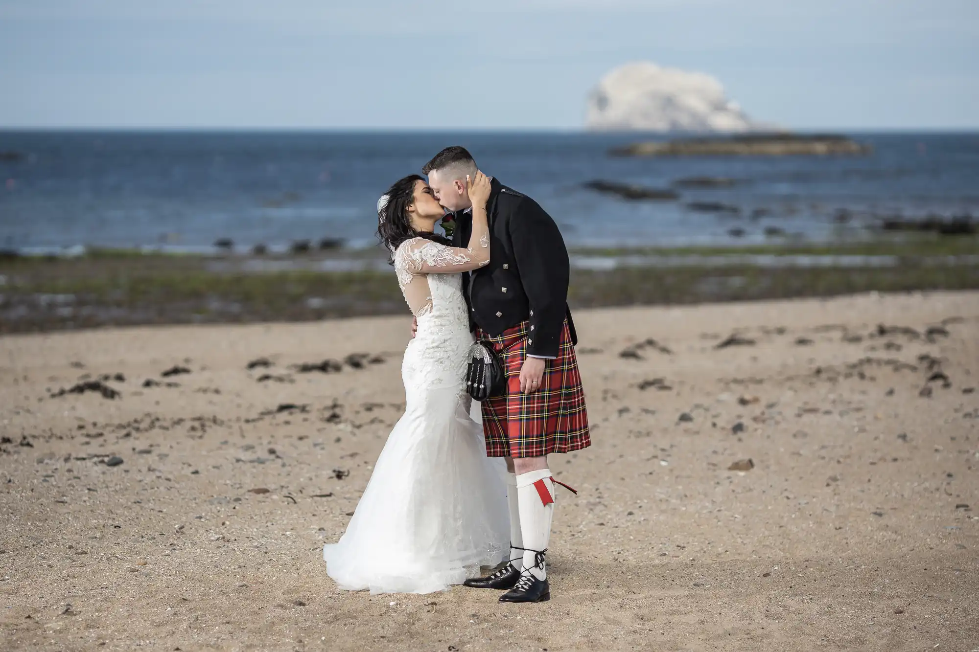 A bride and groom kissing on a beach, with the groom wearing a kilt and the ocean and a rocky island in the background.